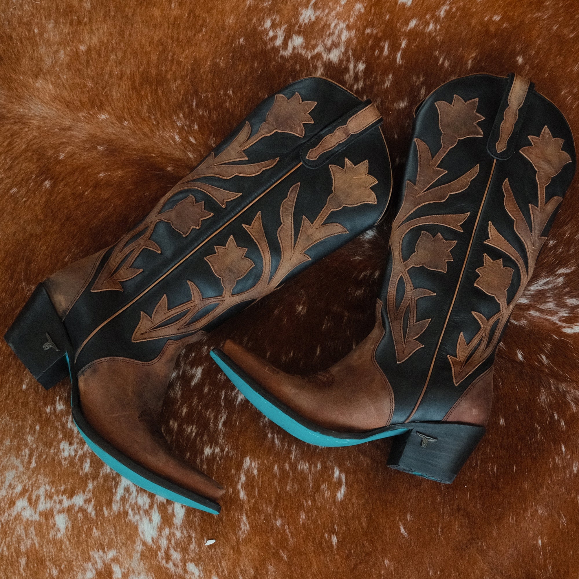 Pair of black and brown cowboy boots with floral inlays and turquoise soles, displayed side by side on a cowhide rug