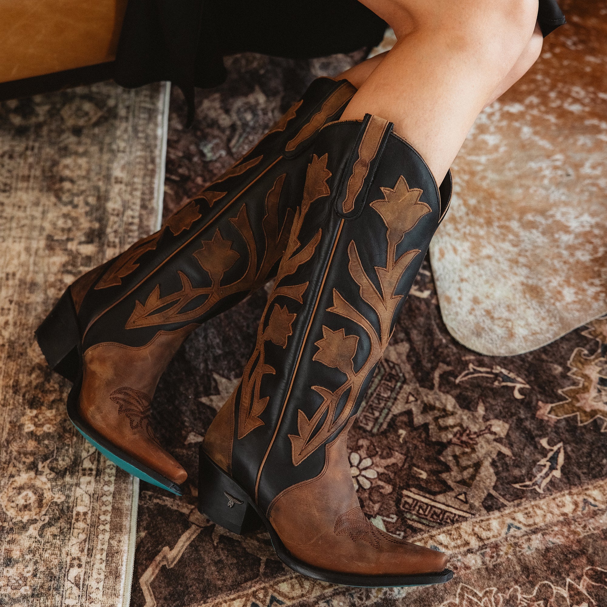 Close-up of black-and-brown cowboy boots with floral inlays and turquoise soles, worn with a black dress, resting on a patterned rug