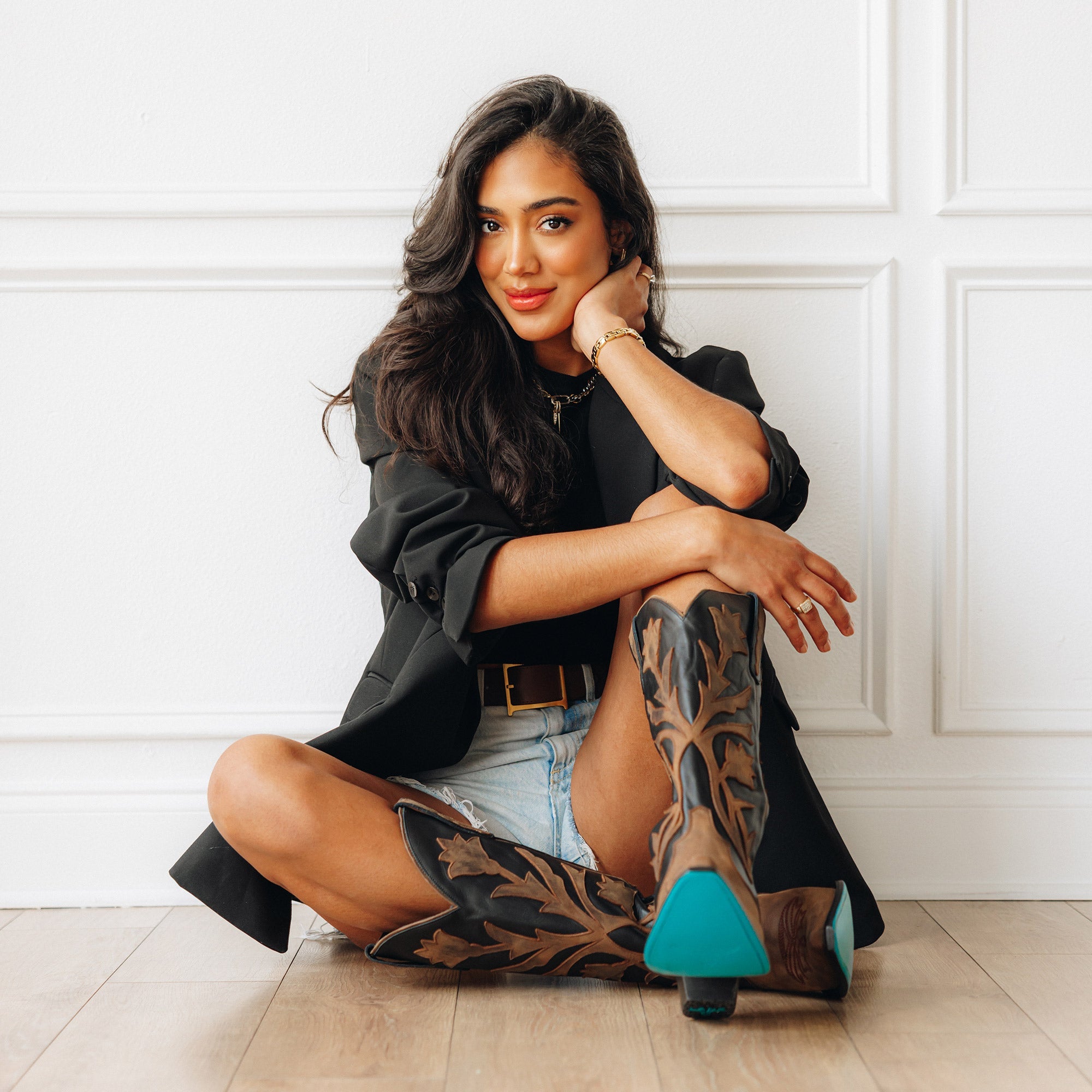 Woman sitting on the floor wearing black and brown cowboy boots with bold leaf inlays and turquoise soles, styled with a black blazer and denim shorts