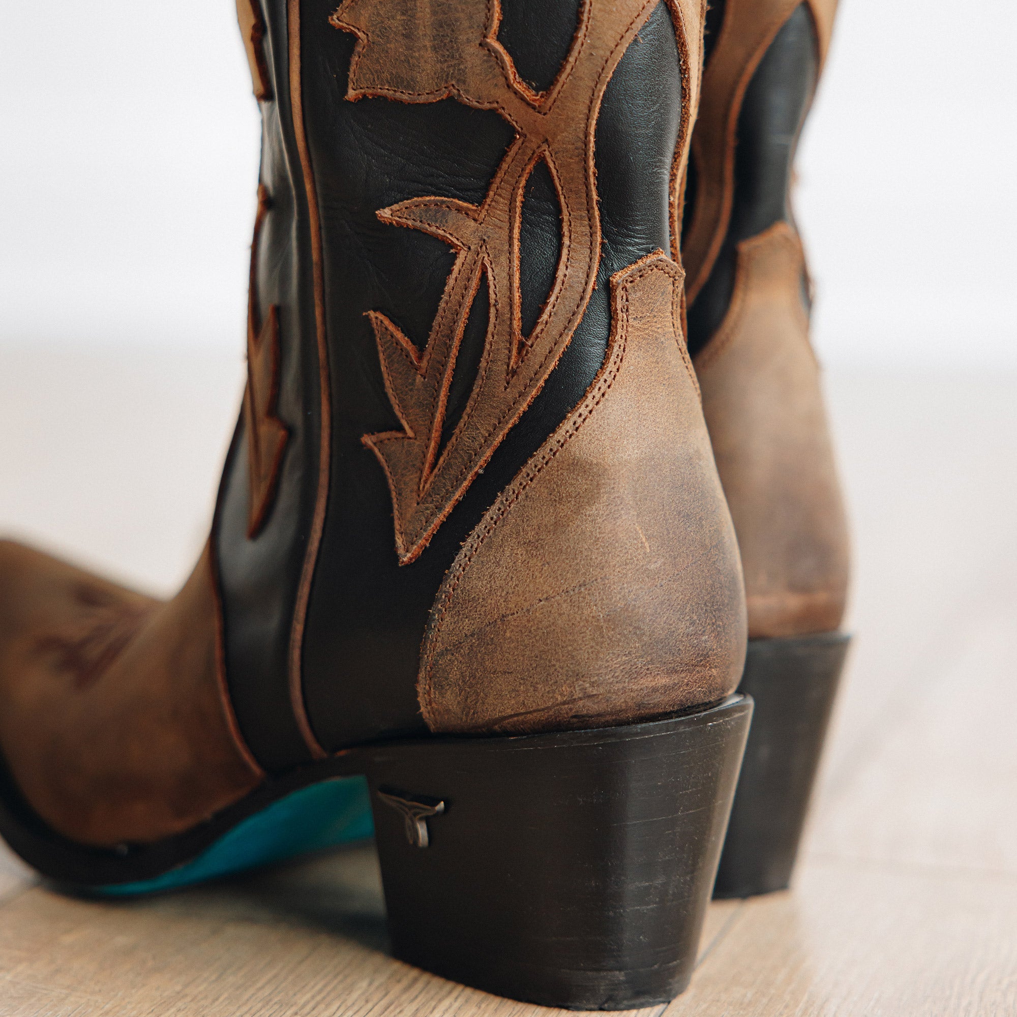 Close-up of the heel and lower shaft of brown and black cowboy boots featuring floral inlays, stacked angled heels, and turquoise soles