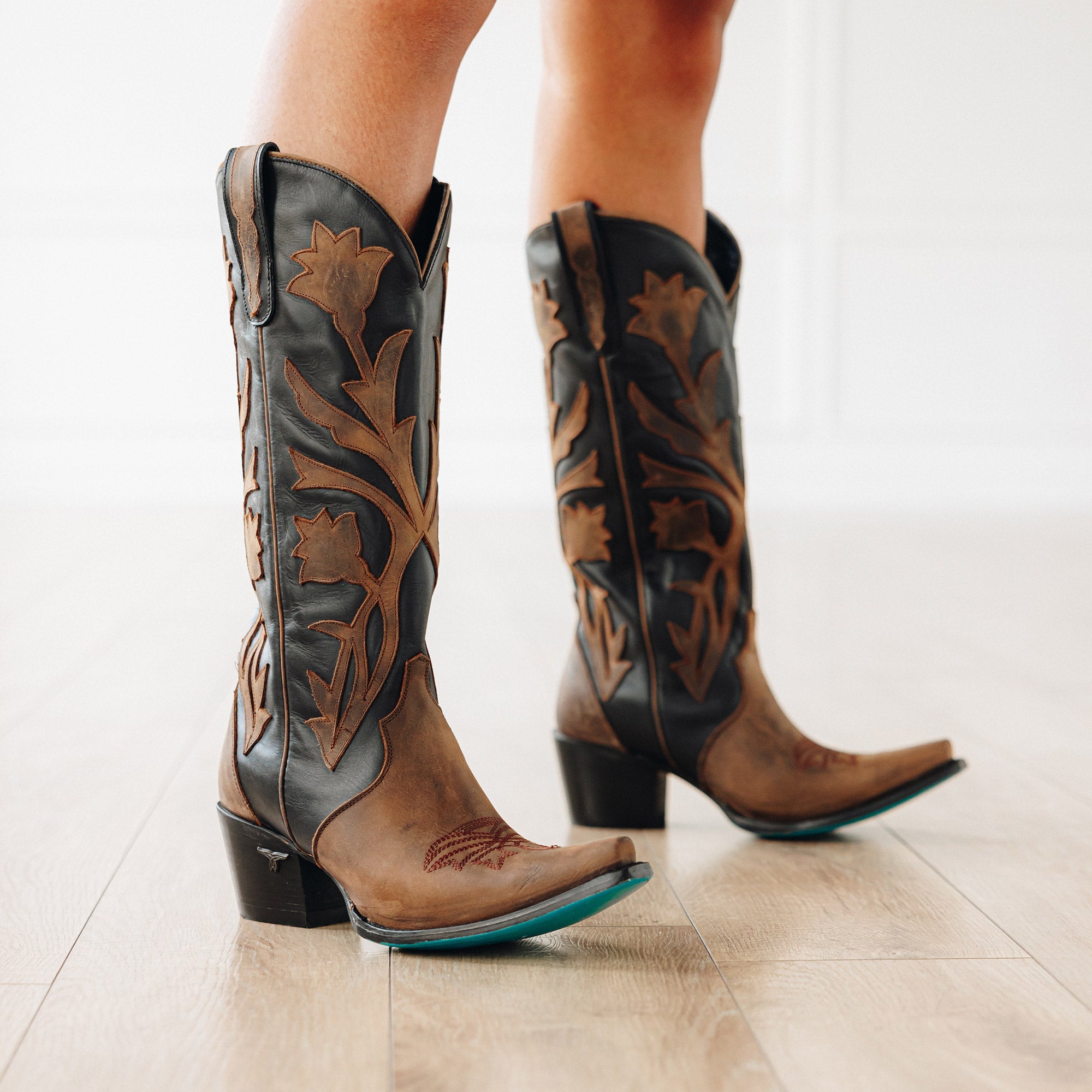 Pair of black and brown cowboy boots with bold floral inlays and turquoise soles, shown being worn on a light wood floor