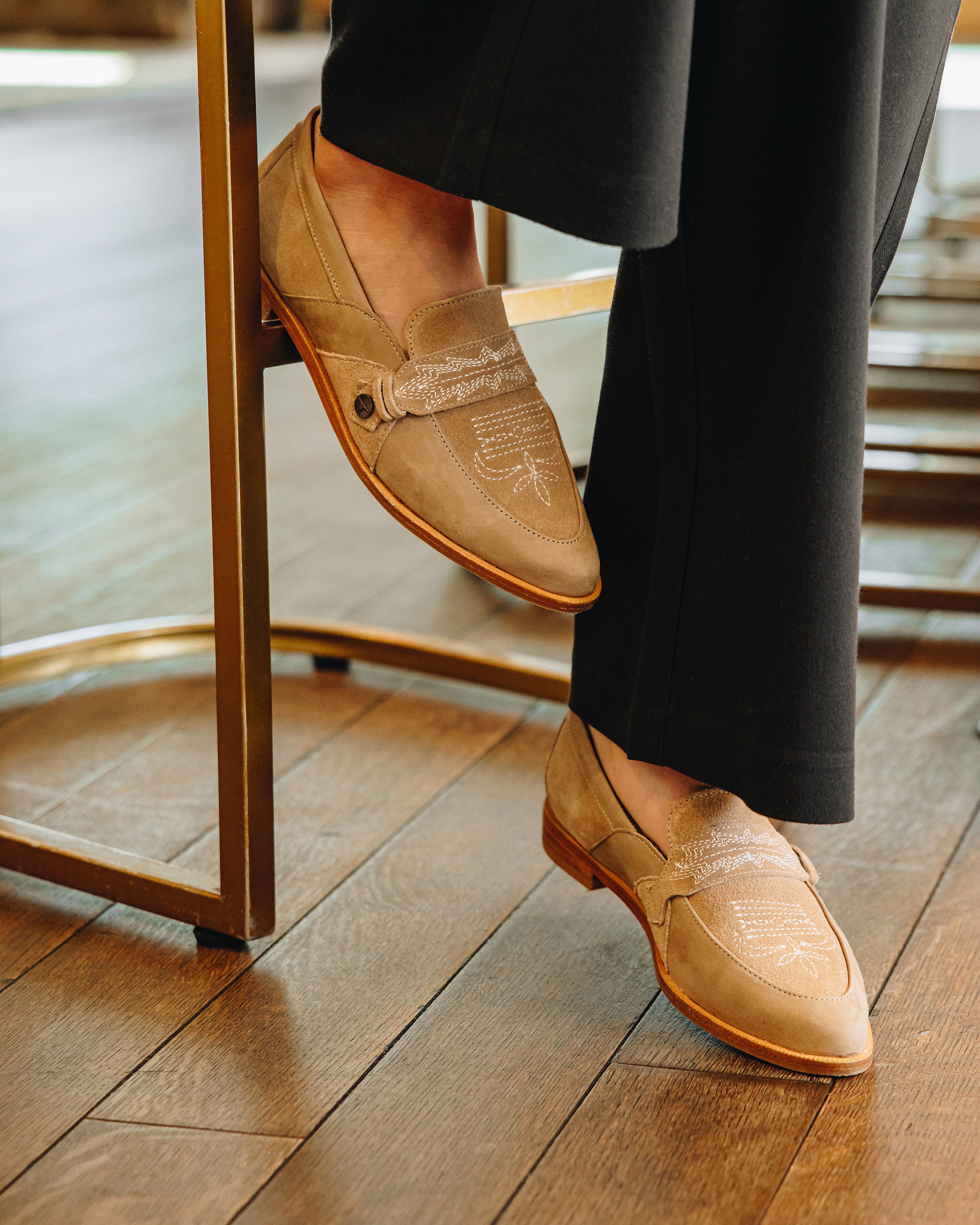 Close-up of tan loafers with decorative stitching and a strap detail, worn with black pants and resting on a brass chair leg over wooden flooring