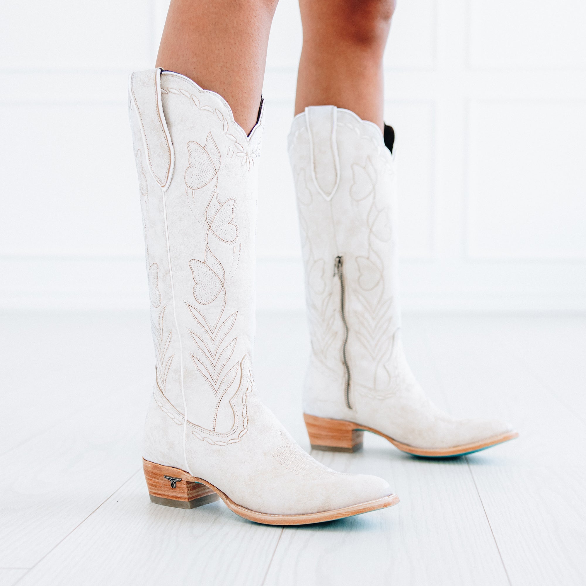 Person wearing tall white cowboy boots with floral stitching, wooden heels, and turquoise soles, standing on a light floor
