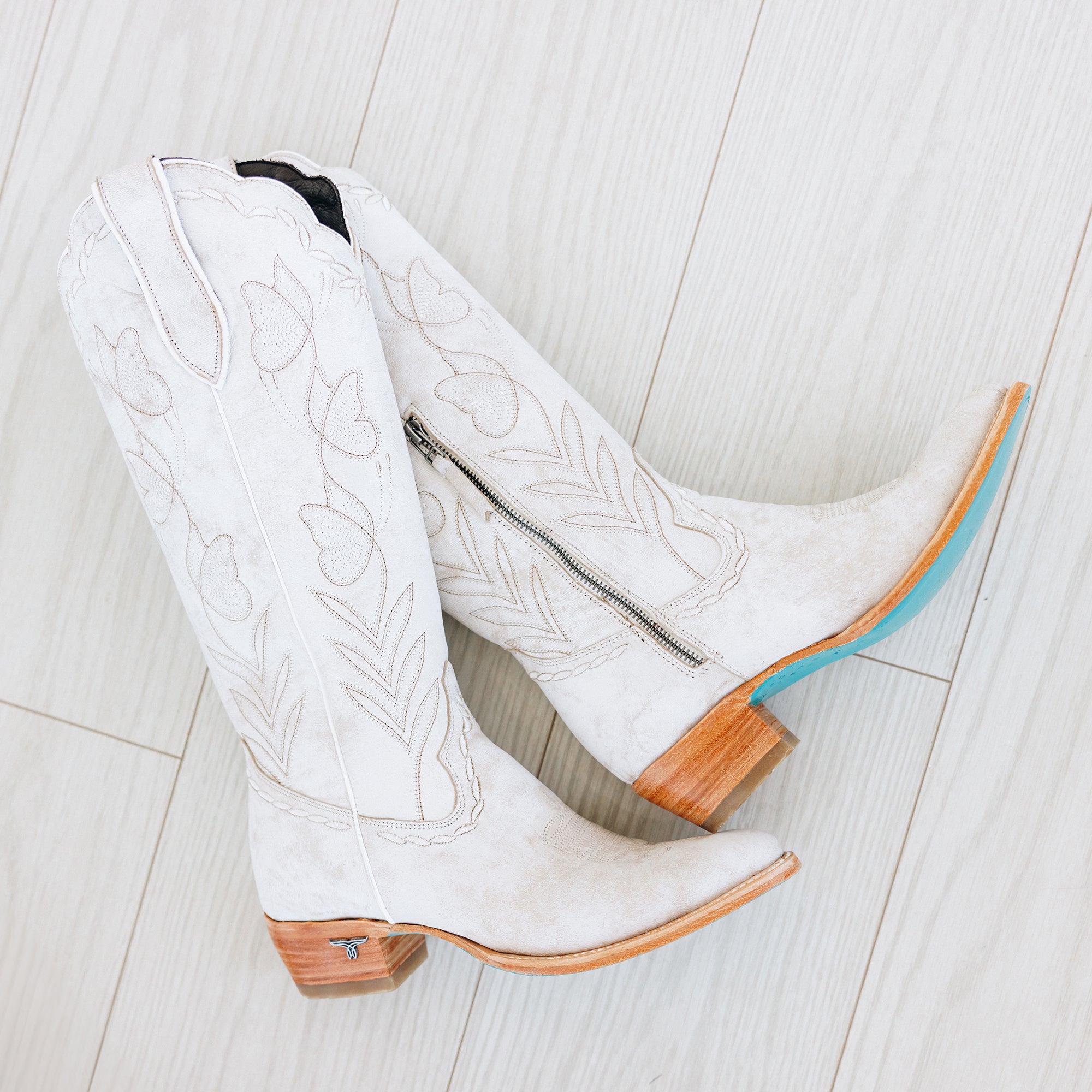 Pair of tall white cowboy boots with floral stitching, wooden heels, turquoise soles, and side zippers, placed on a light wood floor