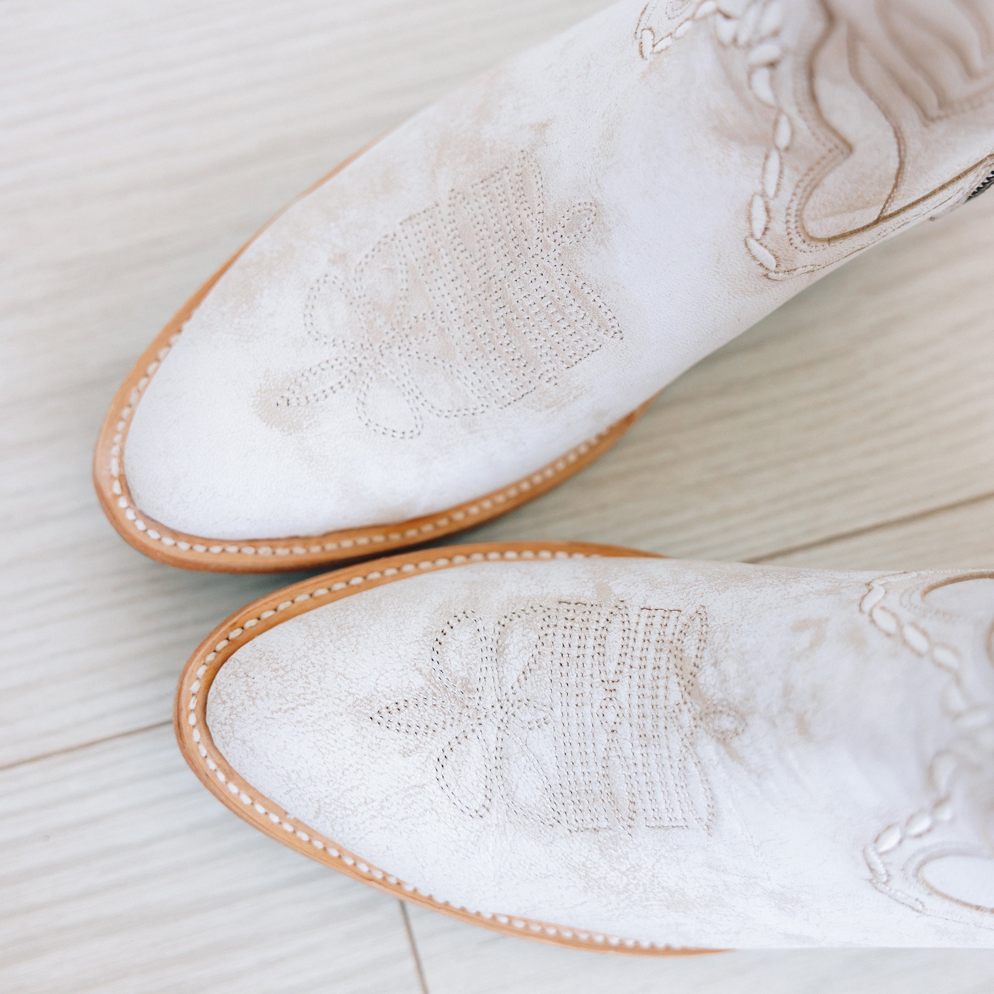 Close-up top view of white cowboy boots with light decorative stitching and tan soles on a light wood floor