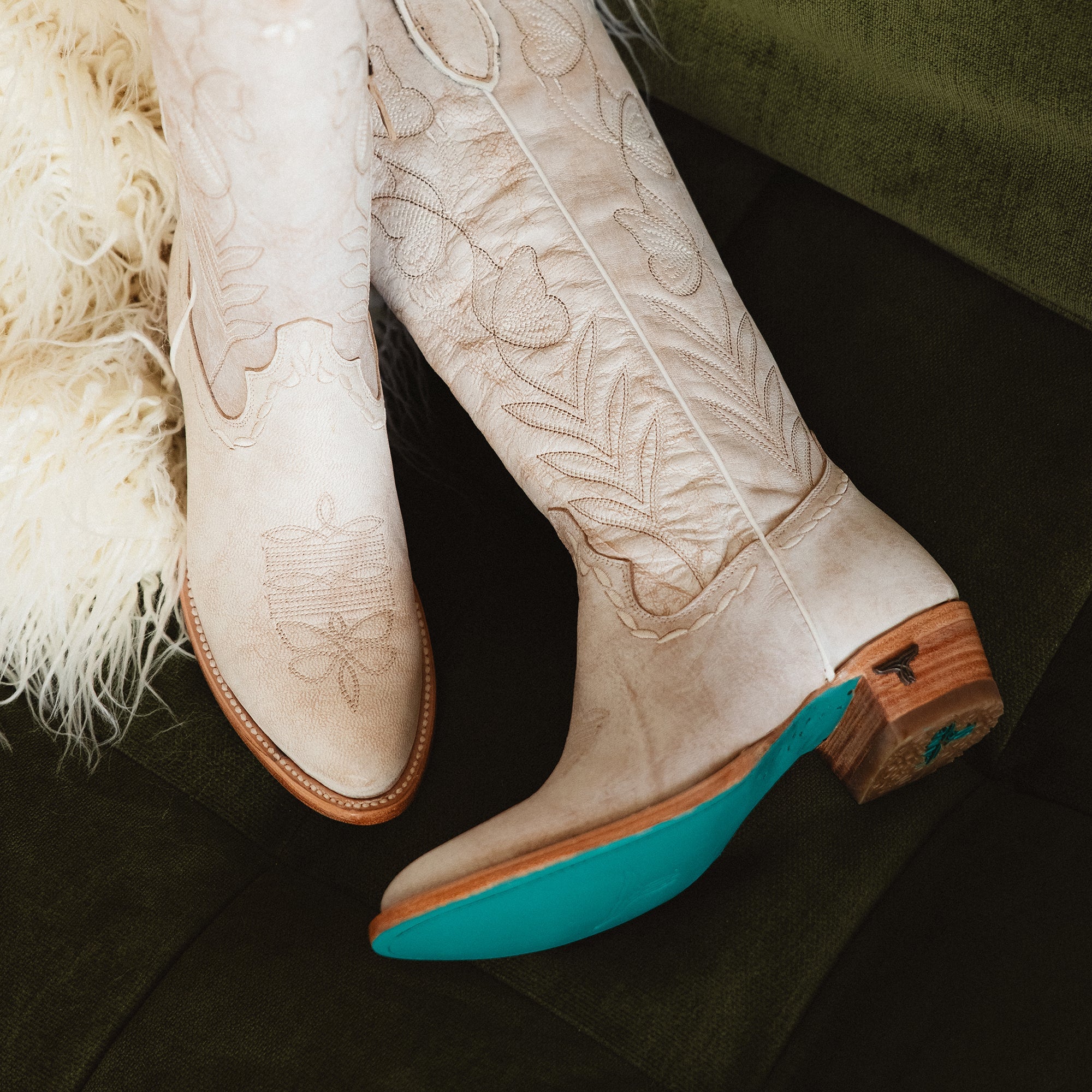 Pair of white cowboy boots with floral stitching, wooden heels, and turquoise soles resting on a green couch next to a fluffy cream throw