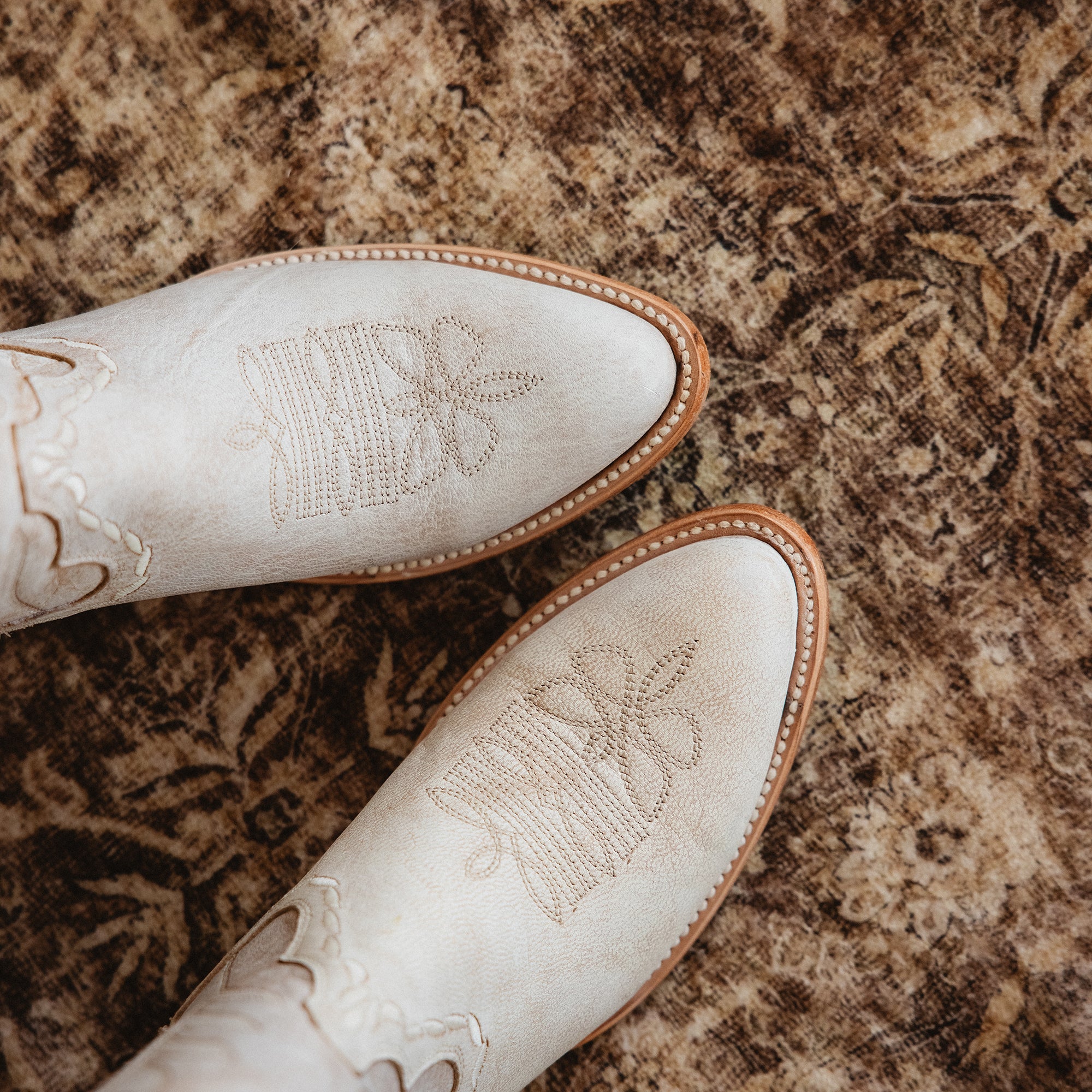 Close-up top view of white cowboy boots with light decorative stitching and tan soles on a patterned rug