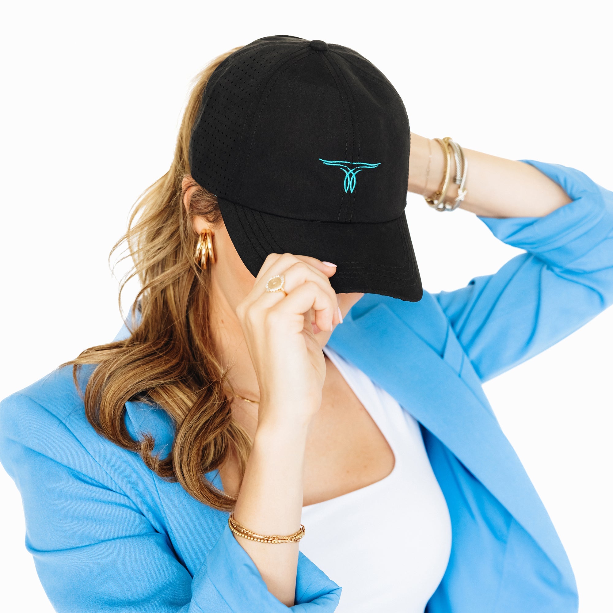 Woman wearing a black baseball cap with turquoise embroidered logo, tilting the brim down with her hand. She has long hair in a ponytail and is dressed in a blue blazer and white shirt, standing against a white background