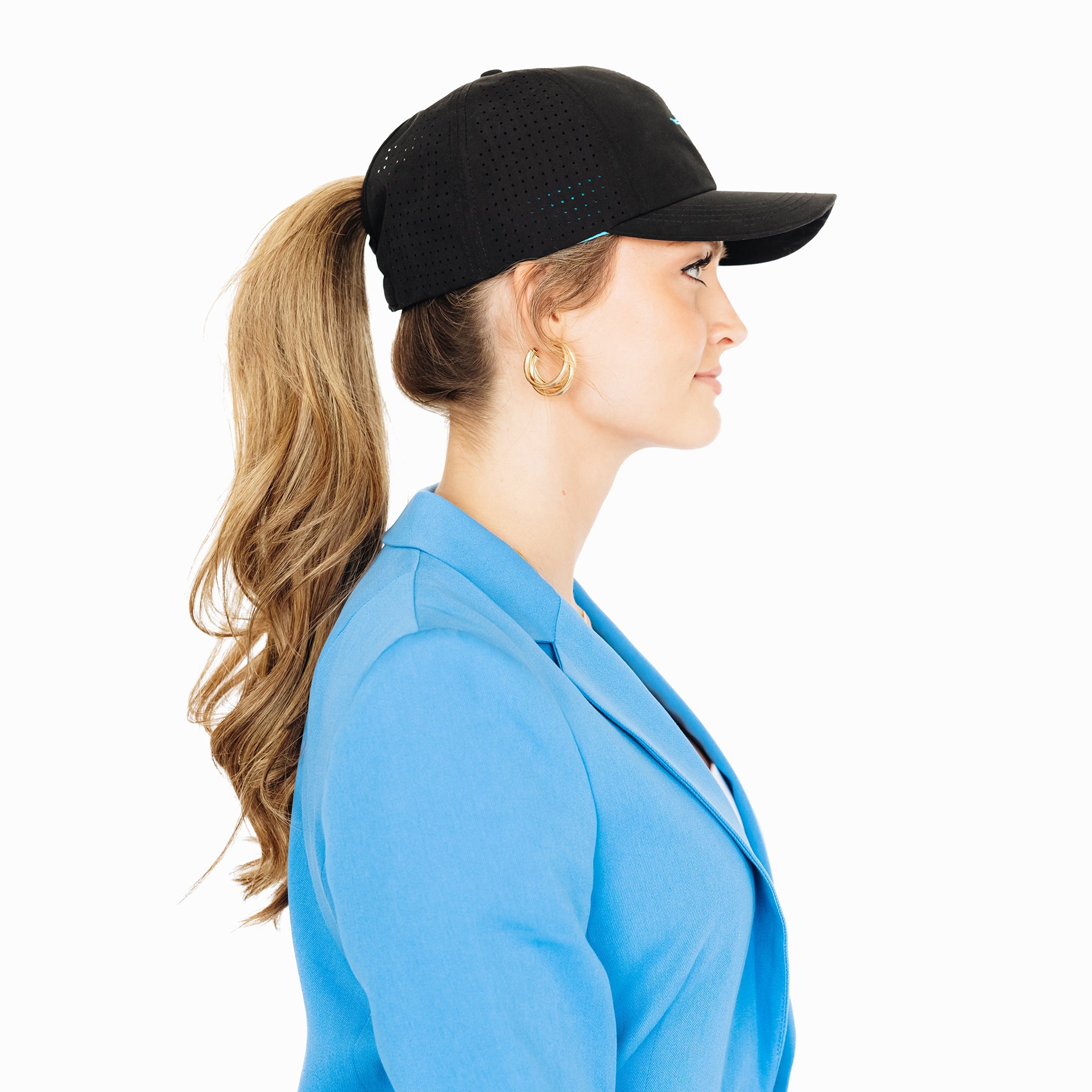 Side profile of a woman wearing a black baseball cap with her ponytail pulled through the back opening. She is dressed in a bright blue blazer and gold hoop earrings, standing against a white background