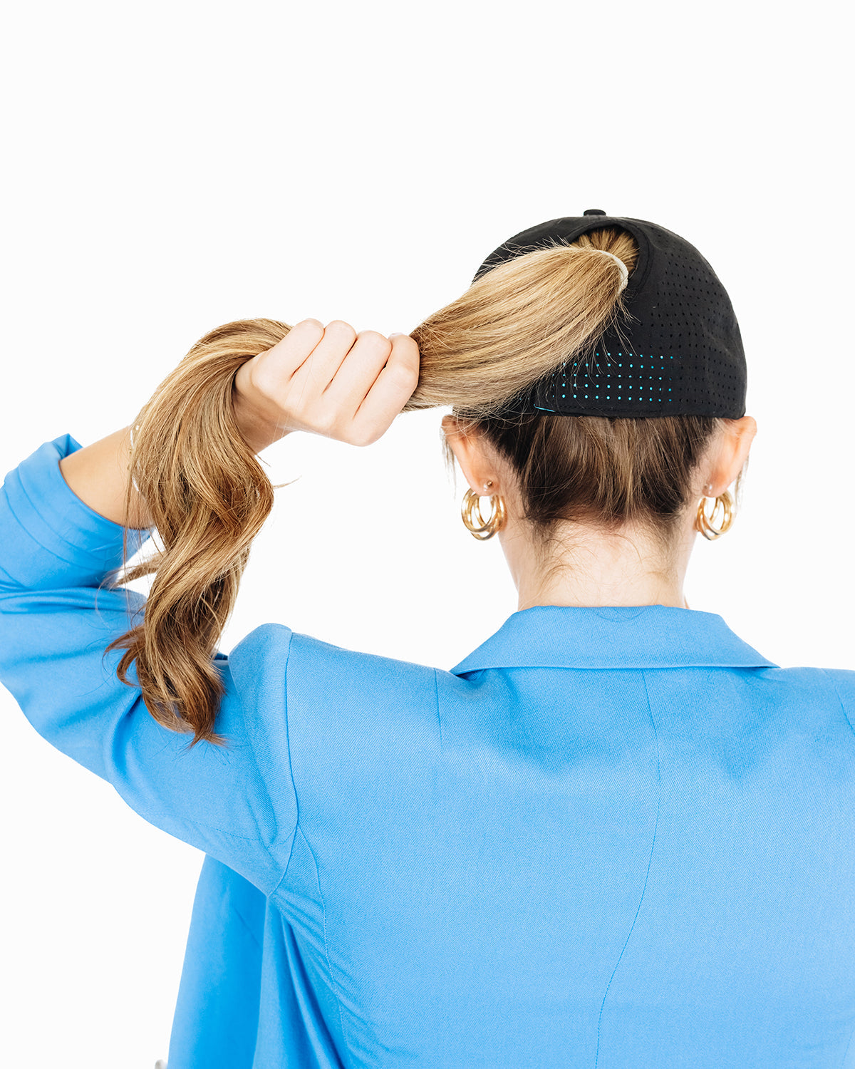Back view of a woman wearing a black perforated baseball cap and a bright blue blazer, holding her ponytail through the hat’s back opening against a white background