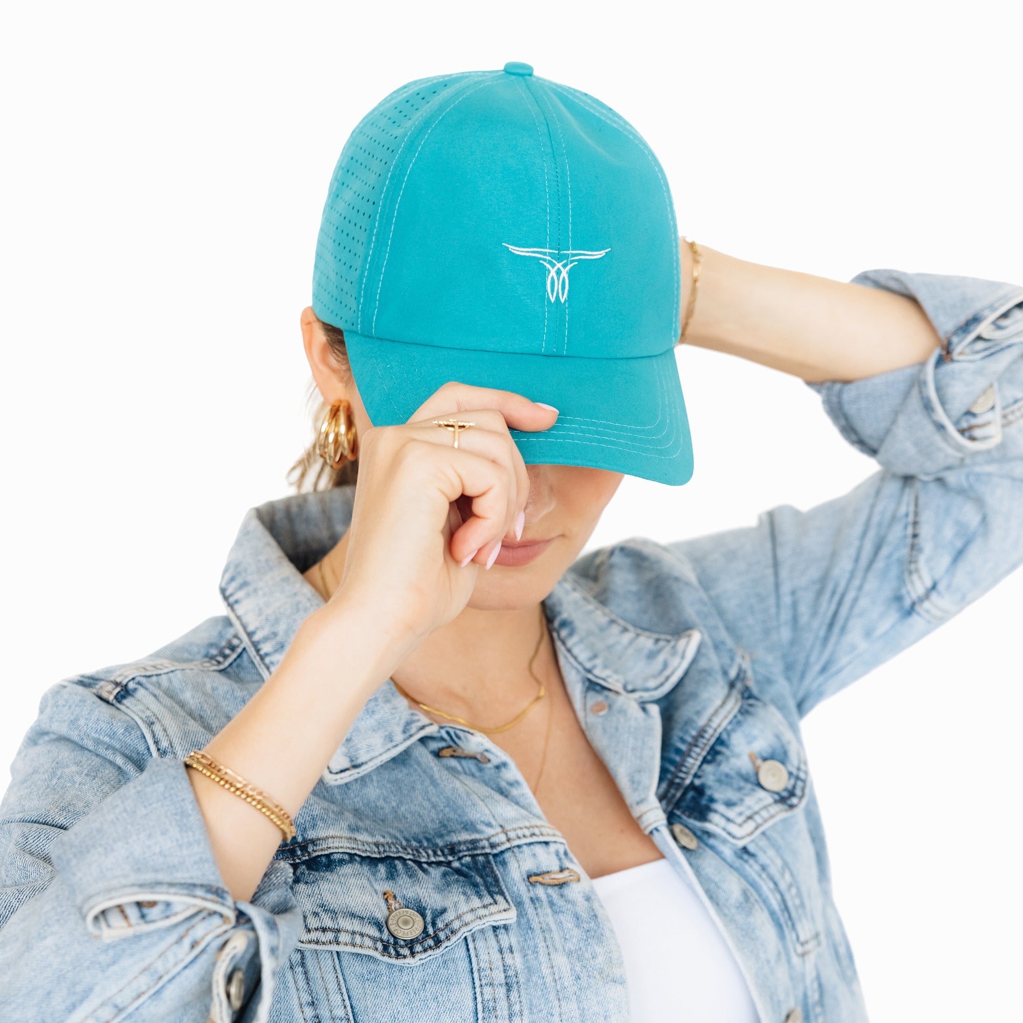 Woman wearing a turquoise baseball cap with a white embroidered logo, tilting the brim down with her hand. She is dressed in a denim jacket and white shirt, standing against a white background