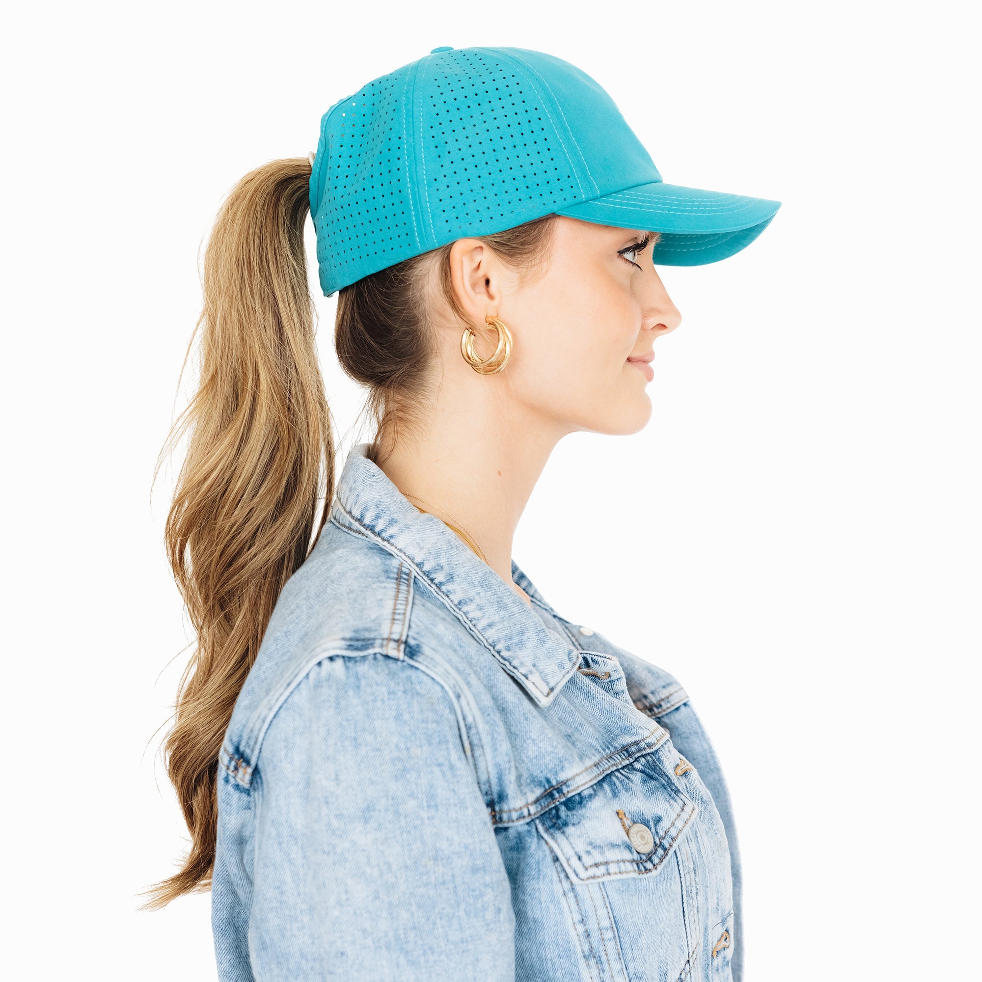 Side profile of a woman wearing a turquoise baseball cap with her ponytail pulled through the back opening. She is dressed in a denim jacket and gold hoop earrings, standing against a white background