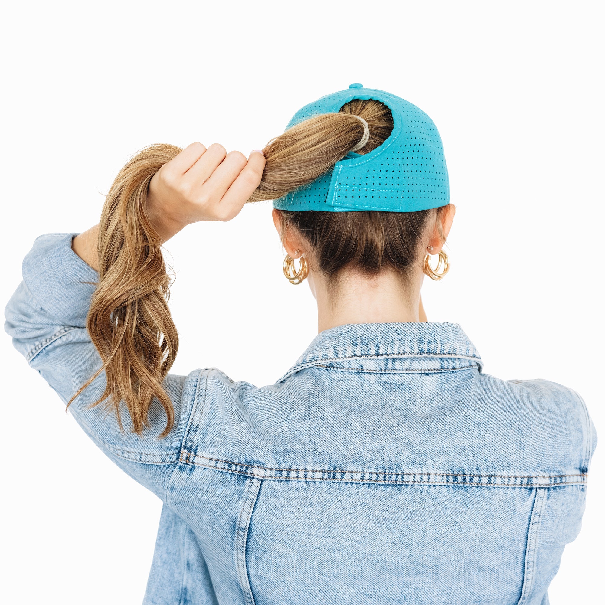 Back view of a woman wearing a turquoise perforated baseball cap and a denim jacket, holding her ponytail through the hat’s back opening against a white background