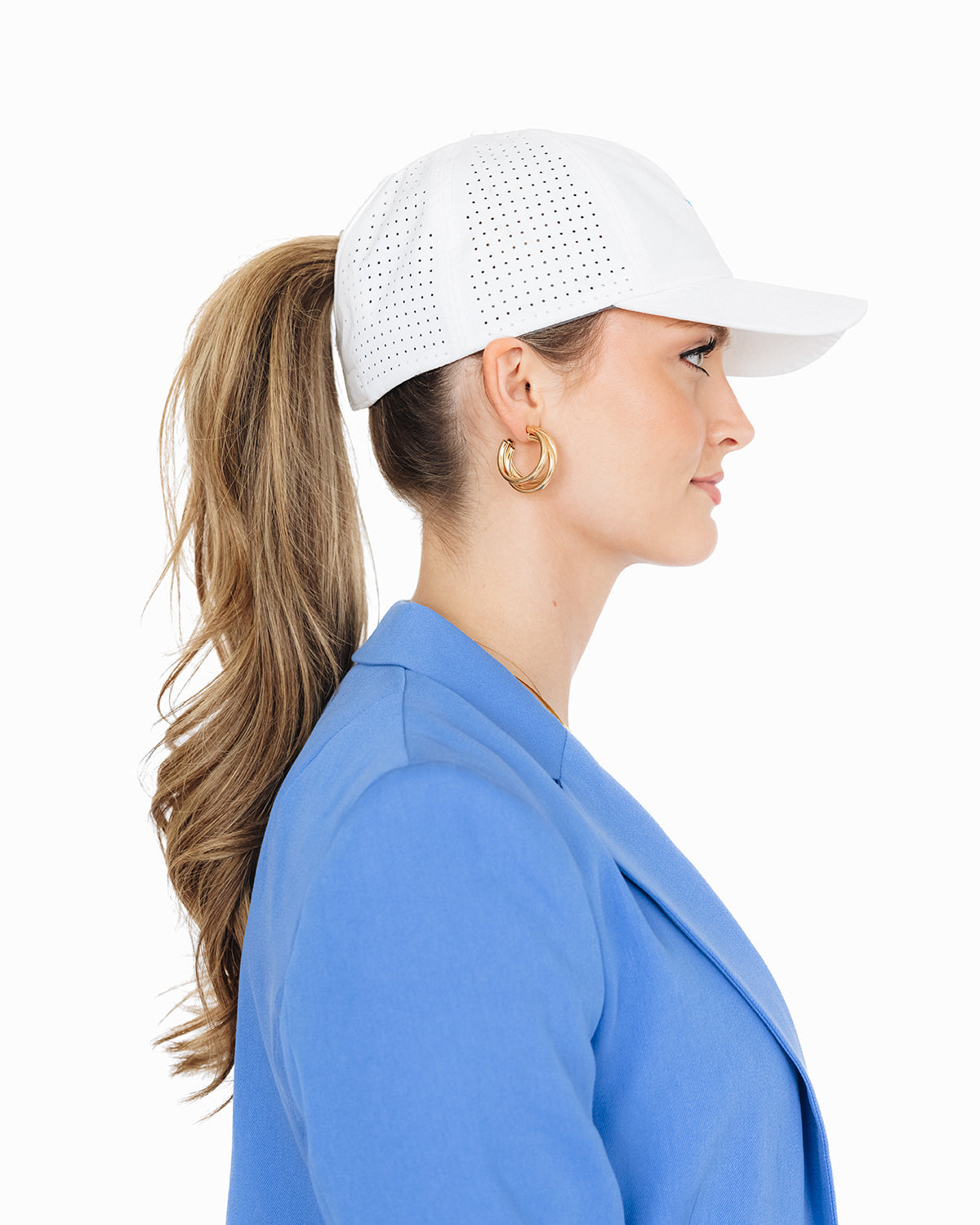 Side profile of a woman wearing a white baseball cap with her ponytail pulled through the back opening. She is dressed in a bright blue blazer and gold hoop earrings, standing against a white background