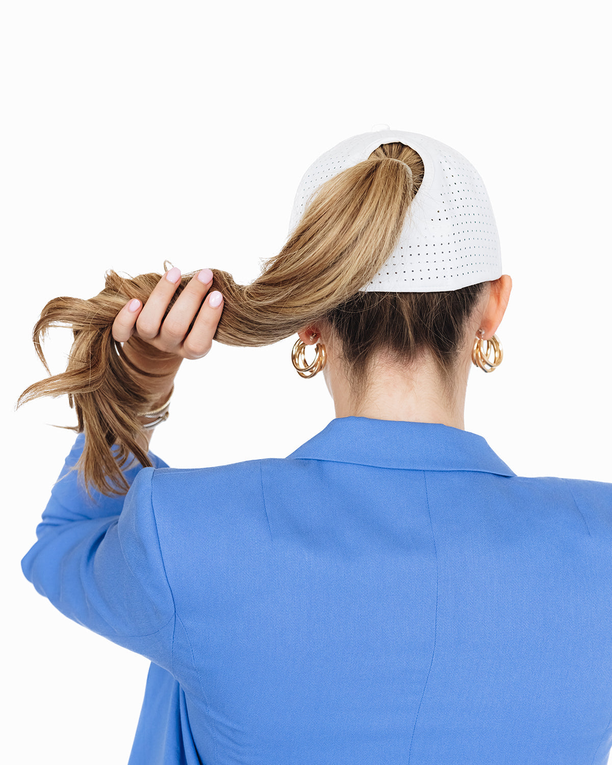 Back view of a woman wearing a white perforated baseball cap and a bright blue blazer, holding her ponytail through the hat’s back opening against a white background