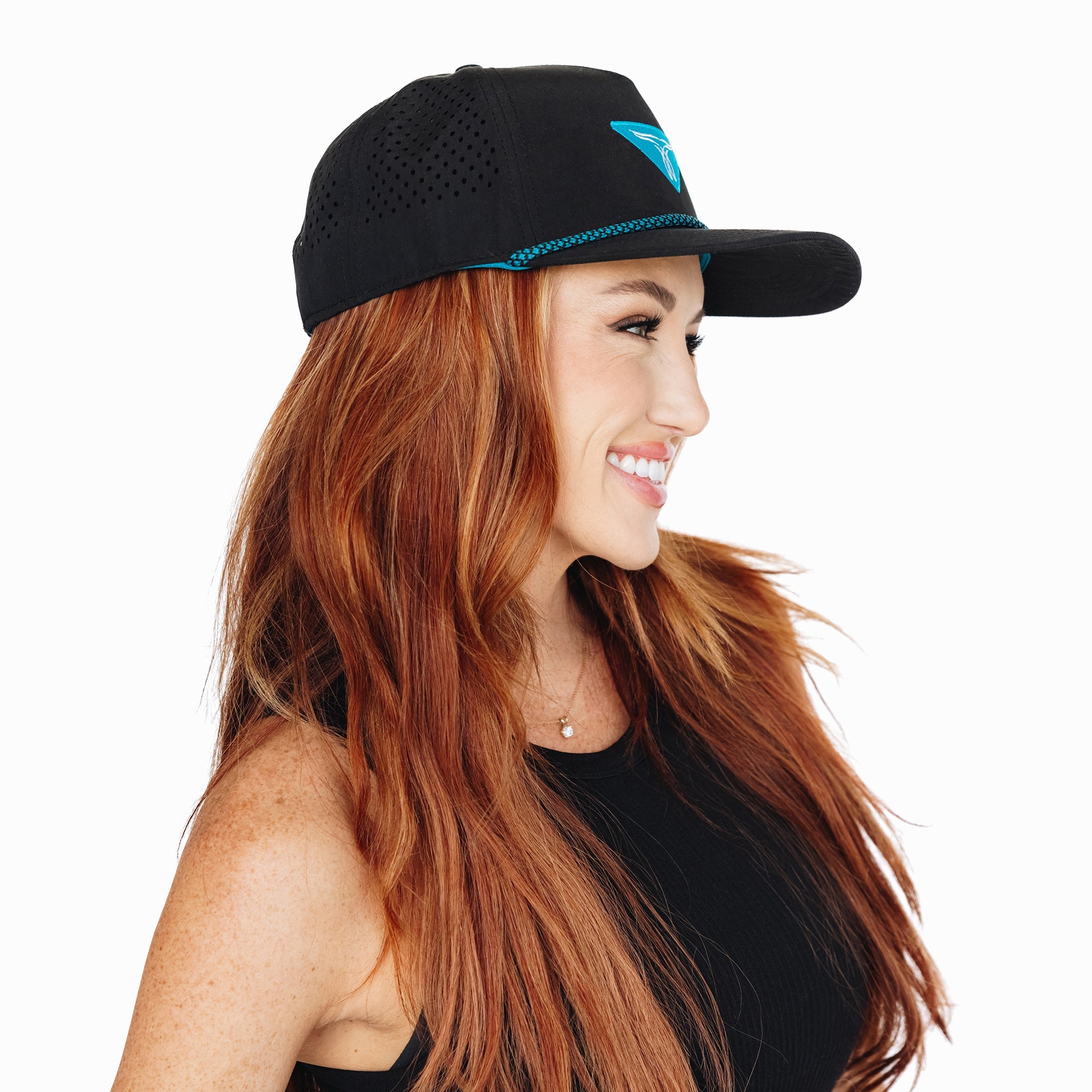 Smiling woman wearing a black perforated snapback hat with a turquoise embroidered logo, paired with a black sleeveless top, facing sideways against a white background