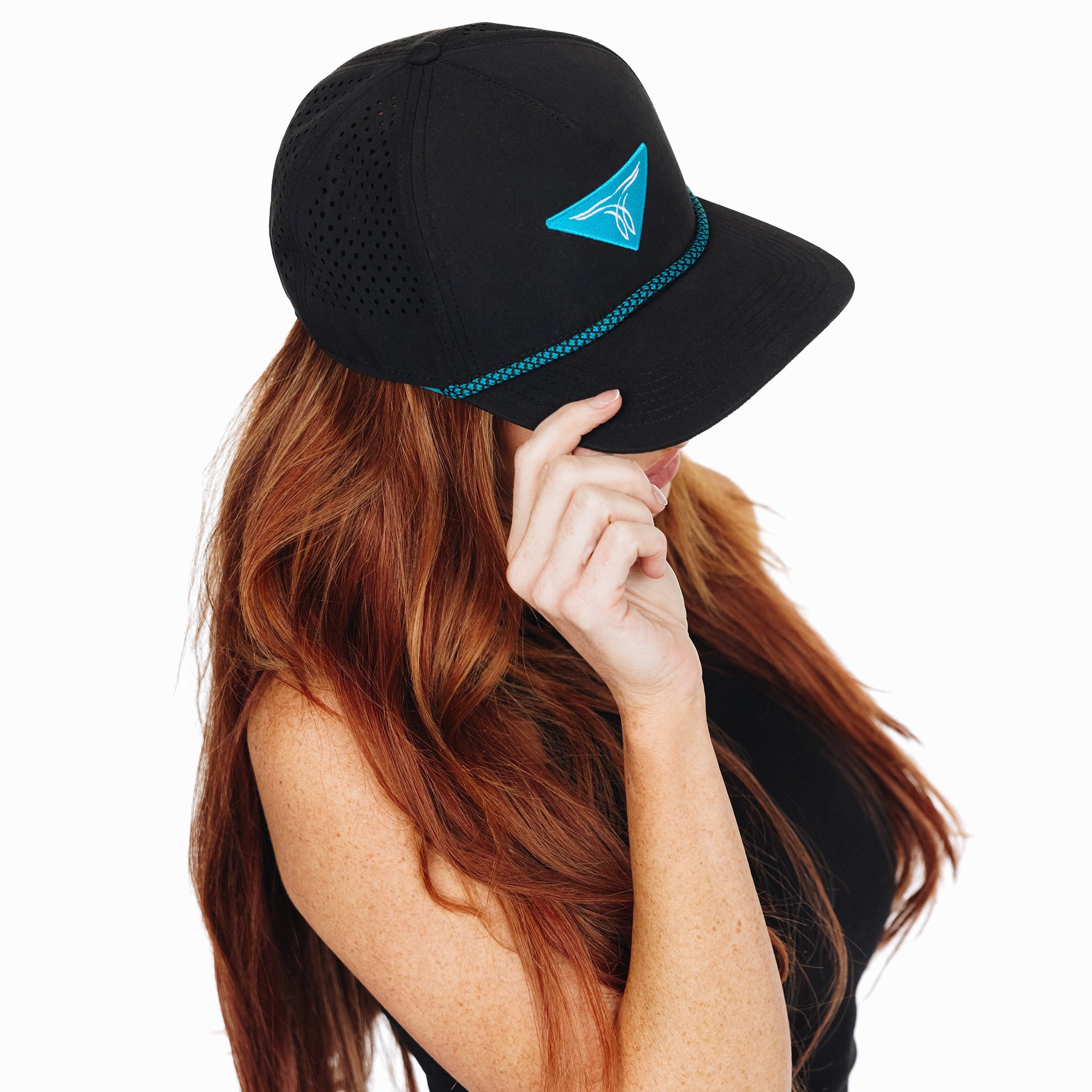 Woman wearing a black baseball cap with turquoise embroidered logo and trim, tilting the brim down with her hand. She has long hair and is dressed in a black top, standing against a white background