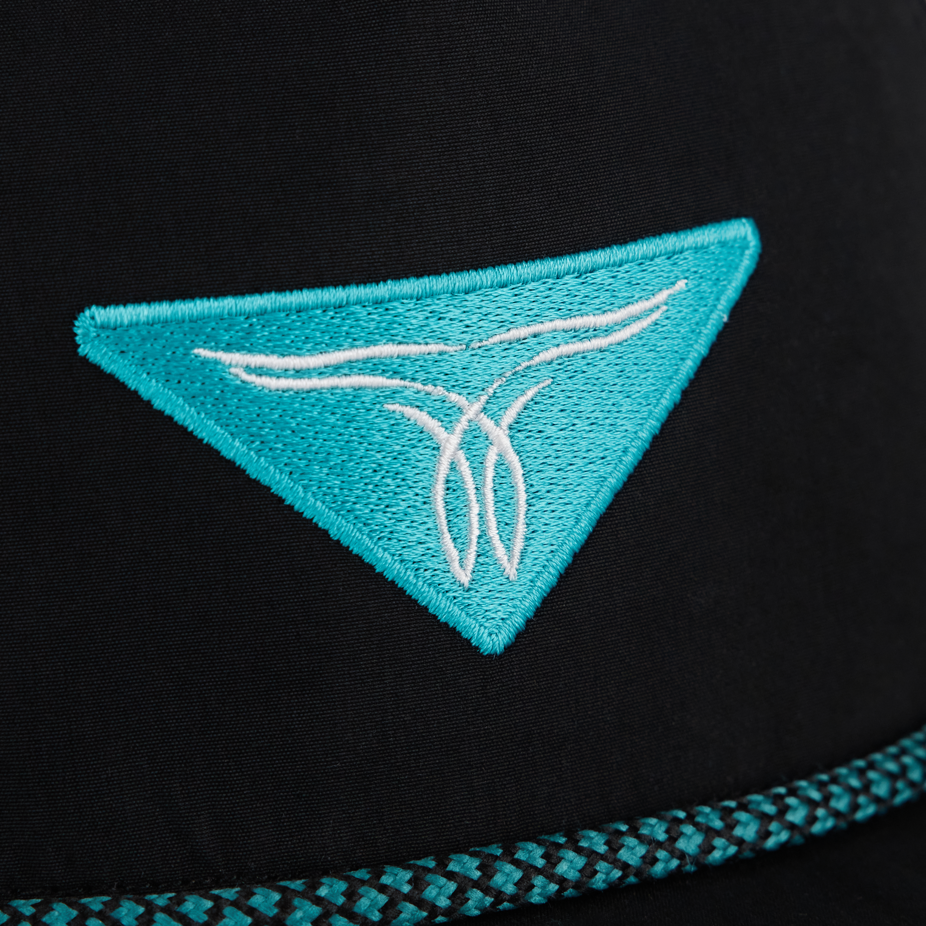 A close-up of a black hat with a turquoise embroidered triangular logo and decorative trim along the edge