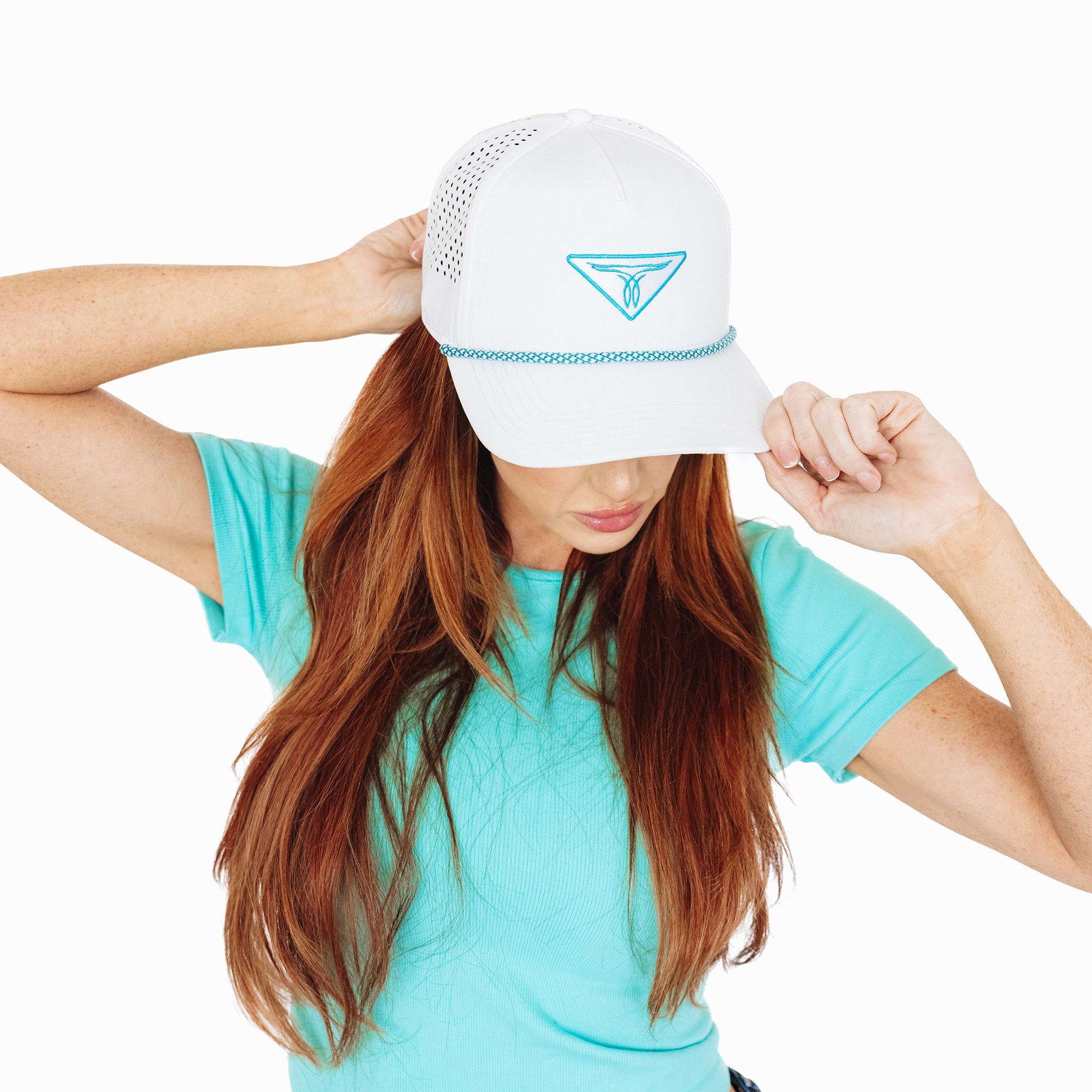Woman wearing a white baseball cap with turquoise embroidered logo and trim, tilting the brim down with her hand. She has long hair and is dressed in a turquoise top, standing against a white background