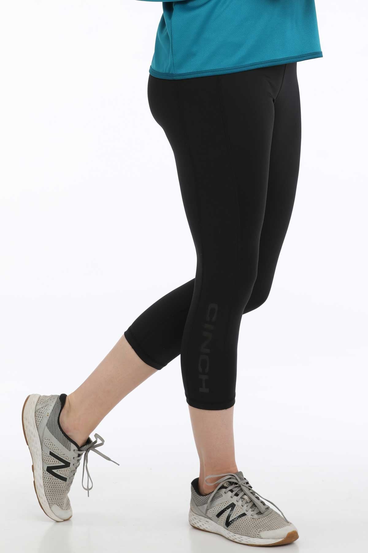Side view of a woman wearing black cropped leggings with sneakers and a teal long-sleeve top displayed on a white background.