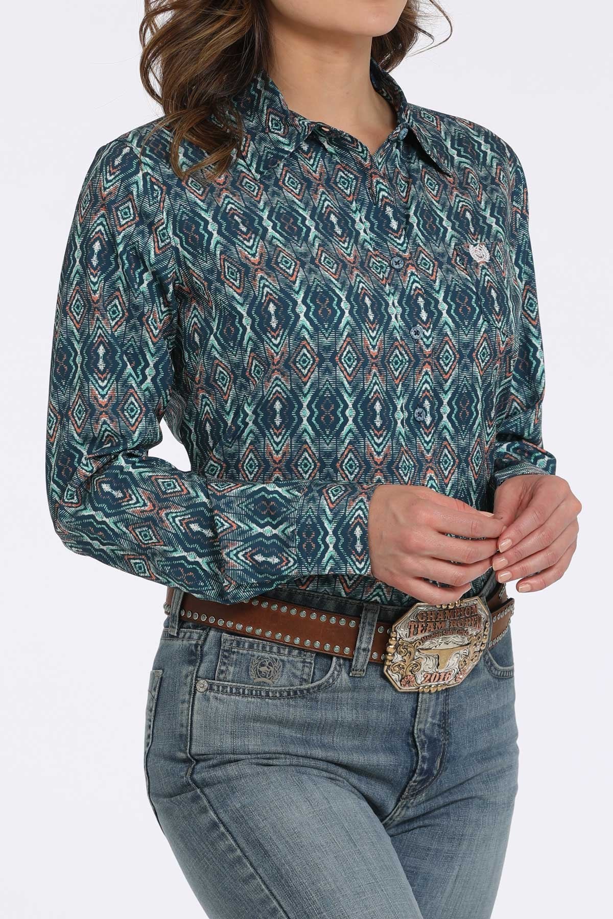 Woman wearing a teal button-down shirt with a geometric diamond pattern, paired with blue jeans and a western belt buckle displayed on a white background