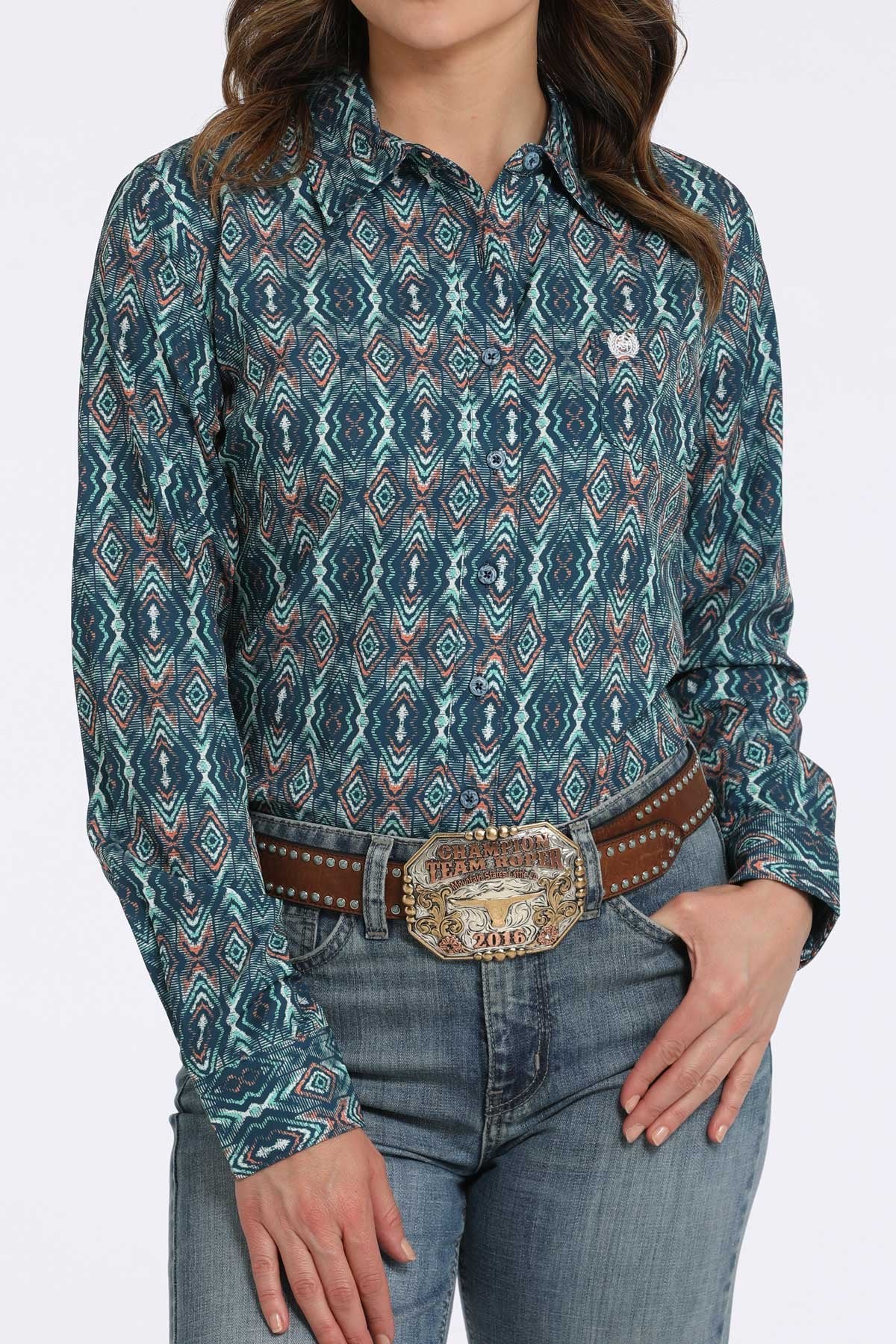 Woman wearing a teal button-down shirt with a geometric diamond pattern, paired with blue jeans and a western belt buckle displayed on a white background