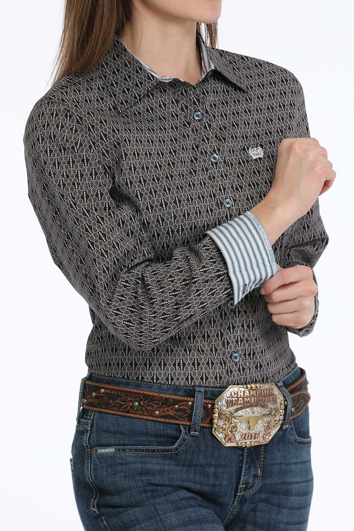 Woman wearing a black button-down long sleeve western shirt with a geometric print, paired with blue jeans and a leather belt with a large buckle. The shirt has a button-down collar, chest logo, and contrasting  fabric inside the cuffs, displayed on a white background