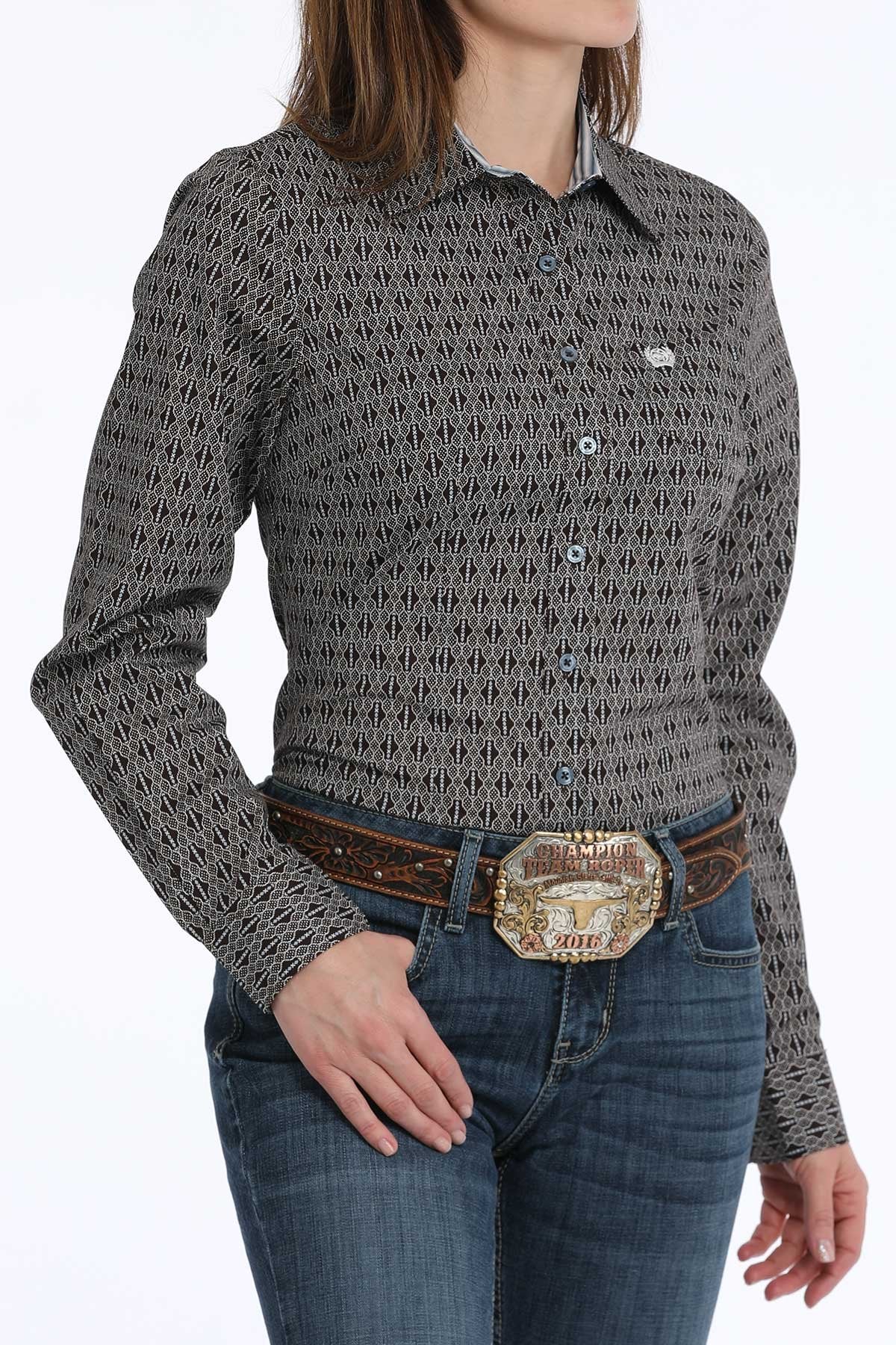 Woman wearing a black button-down long sleeve western shirt with a geometric print, paired with blue jeans and a leather belt with a large buckle. The shirt has a button-down collar, chest logo and is displayed on a white background