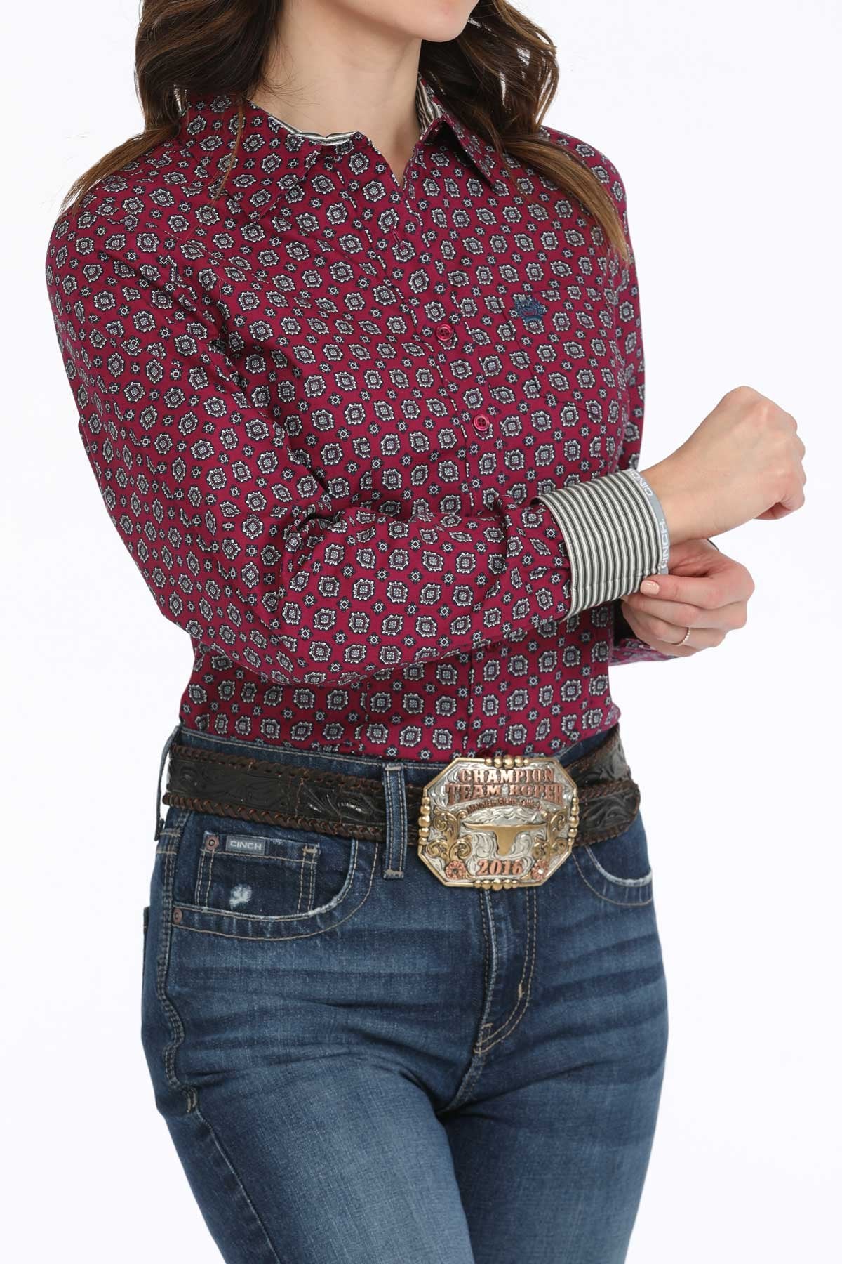 A woman wearing a maroon patterned long-sleeve button-up shirt with contrasting cuffs, dark jeans, and a large belt buckle stands with her arms slightly crossed