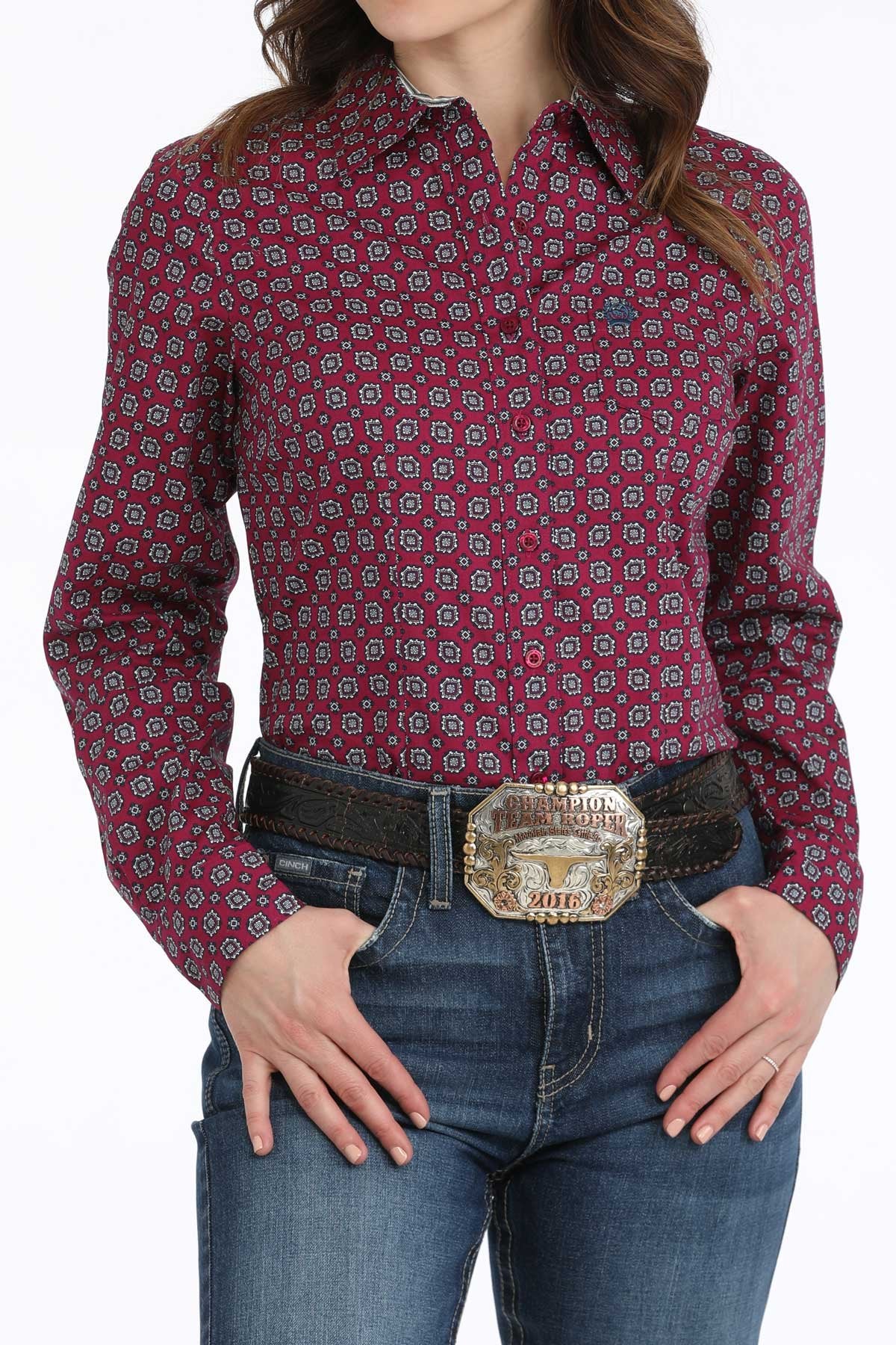 A woman wearing a maroon patterned long-sleeve button-up shirt, dark jeans, and a large belt buckle standing against a white background