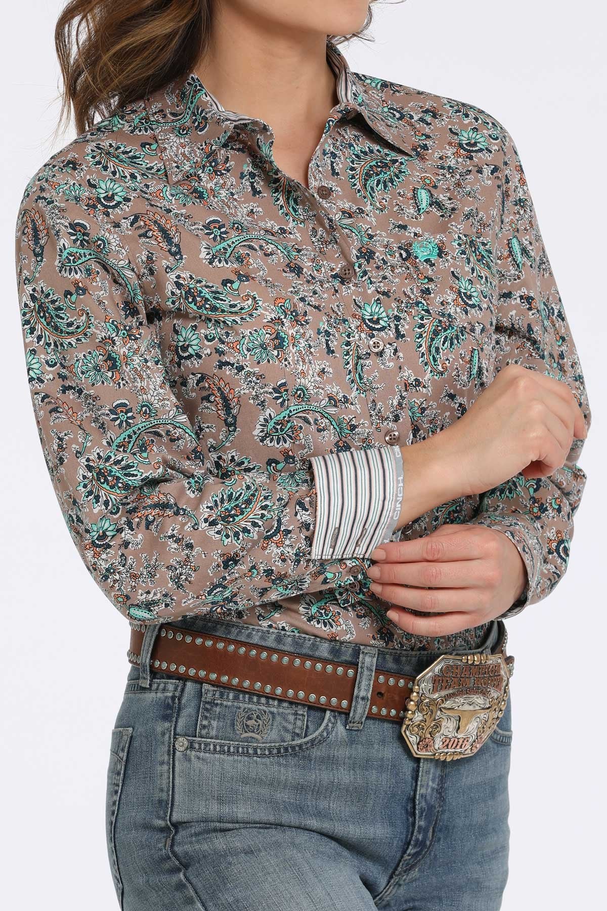 Woman wearing a tan button-down shirt with a teal and black floral paisley pattern, showing contrasting striped fabric on the inside cuff  paired with blue jeans and a western belt buckle displayed on a white background.