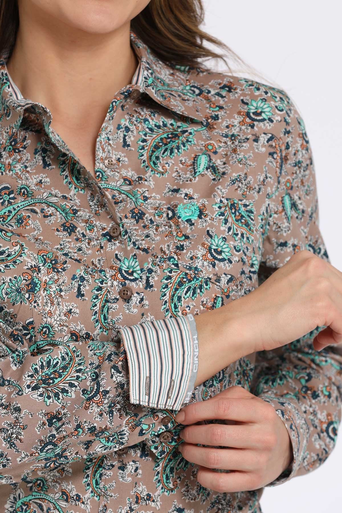 Close-up view of a woman wearing a tan button-down shirt with a teal and black floral paisley pattern, showing contrasting striped fabric on the inside cuff displayed on a white background.