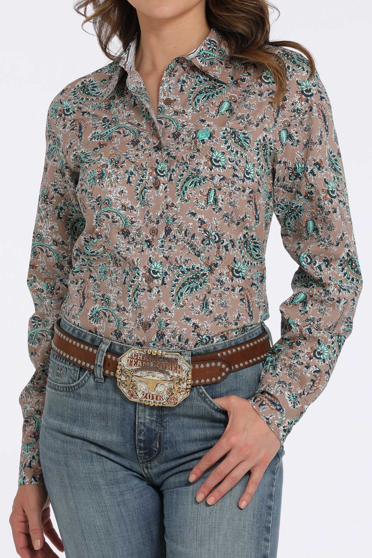 Woman wearing a tan button-down shirt with a teal and black floral paisley pattern, paired with blue jeans and a western belt buckle displayed on a white background.
