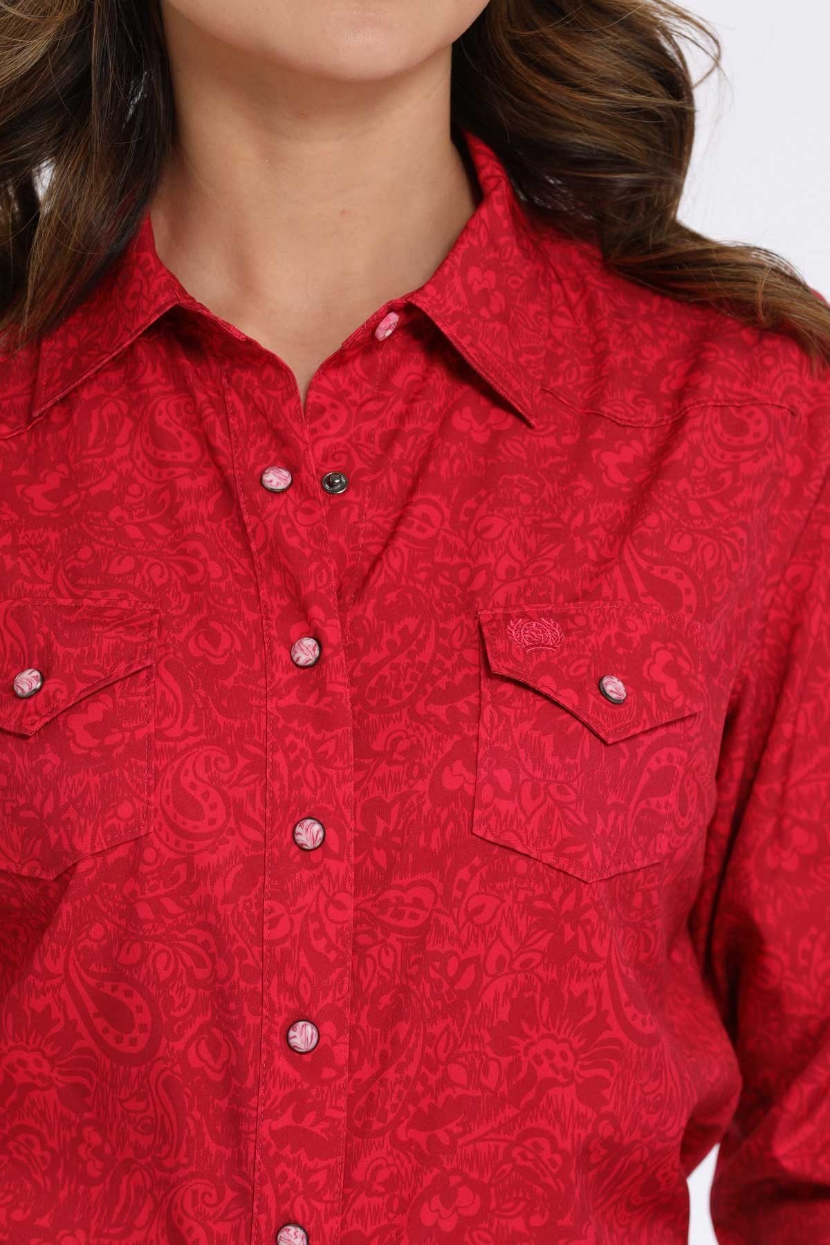 Close-up of a woman wearing a pink long-sleeve button-down shirt with subtle paisley pattern and western-style chest pockets displayed on a white background.