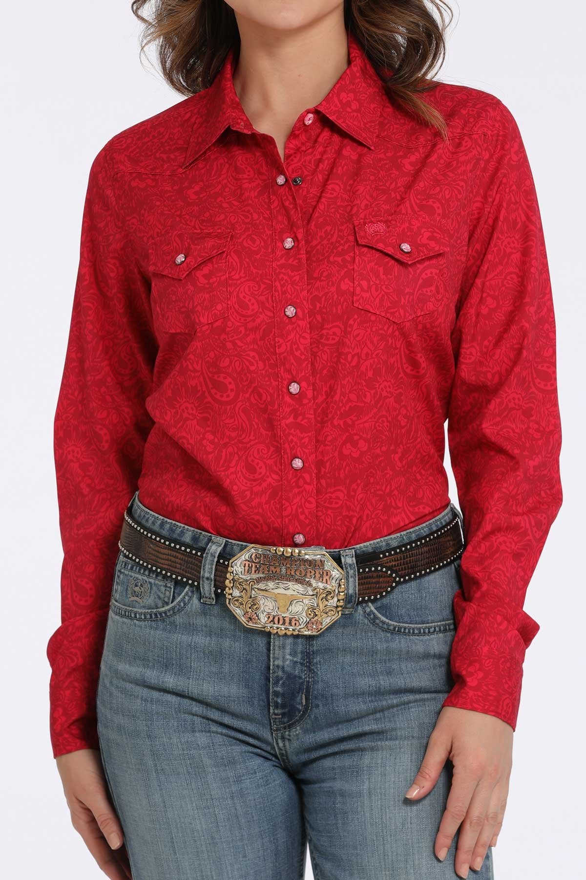 Woman wearing a pink long-sleeve button-down shirt with subtle paisley pattern and western-style chest pockets, paired with blue jeans and a western belt buckle displayed on a white background.