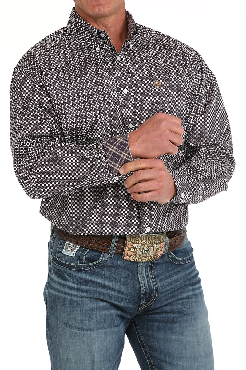 Man wearing a black button-down long sleeve western shirt with a white geometric print, paired with blue jeans and a tooled leather belt with a large buckle. The shirt has a button-down collar, chest logo, and contrasting plaid fabric inside the cuffs, displayed on a white background
