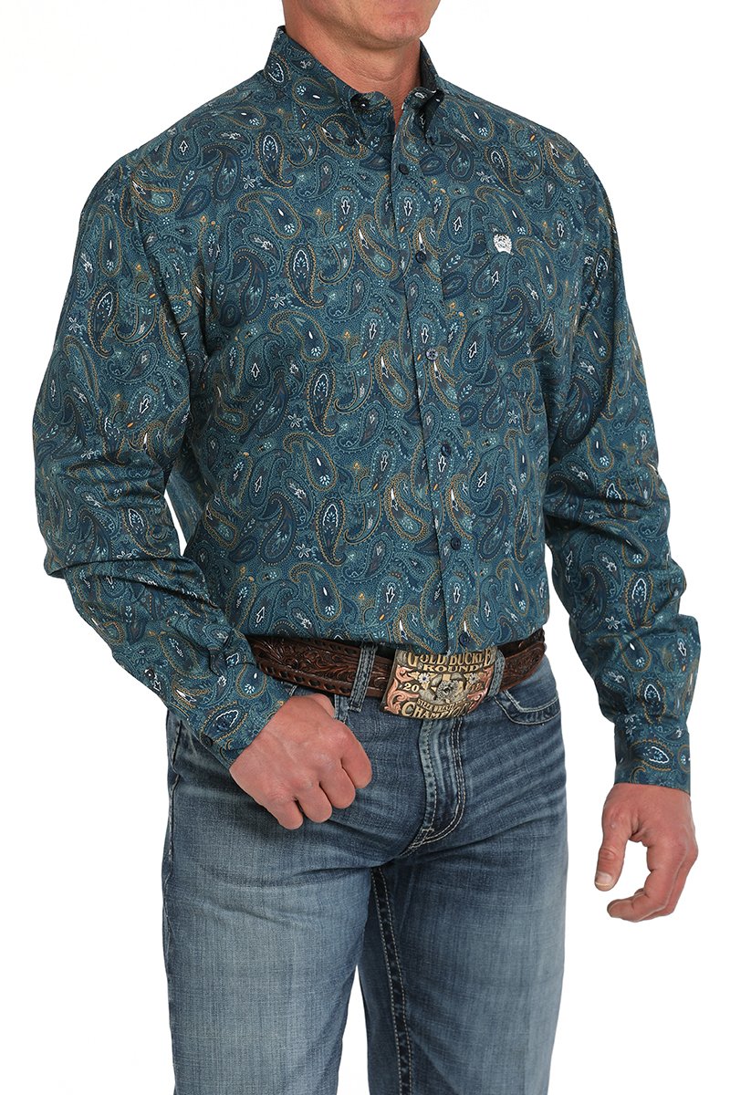 Man wearing a dark teal paisley patterned button-down shirt tucked into blue jeans with a western belt buckle displayed on a white background.