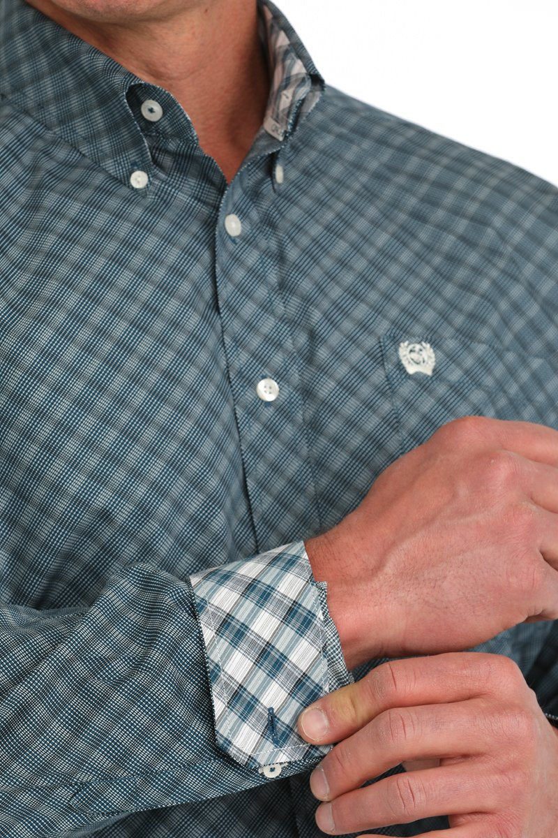 Close-up of a man wearing a teal patterned button-down shirt with rolled sleeves showing a contrasting plaid cuff displayed on a white background.