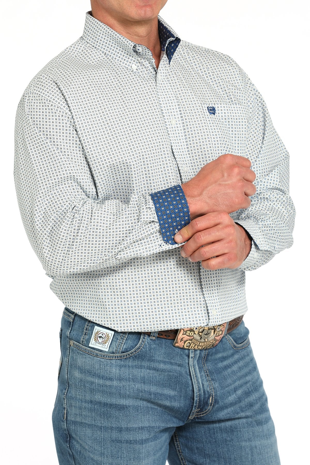 Side angled view of a man wearing a white long-sleeve button-down shirt with a blue geometric print pattern and contrasting cuff detail, paired with blue jeans and a western belt buckle displayed on a white background