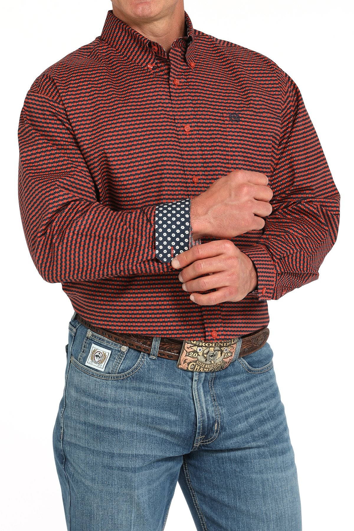 Man wearing a red patterned button-down shirt with contrasting navy floral print on the inside cuff tucked into blue jeans with a western belt buckle displayed on a white background.