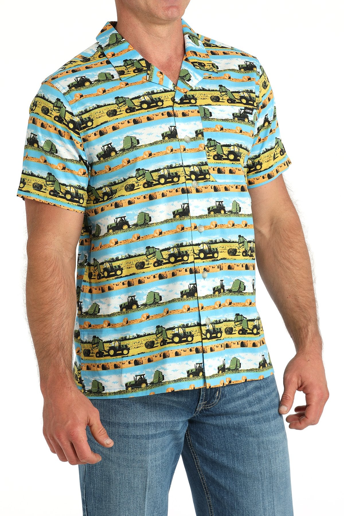 Side angled view of a man wearing a blue short-sleeve button-down shirt with a scenic truck and farm print, paired with blue jeans displayed on a white background