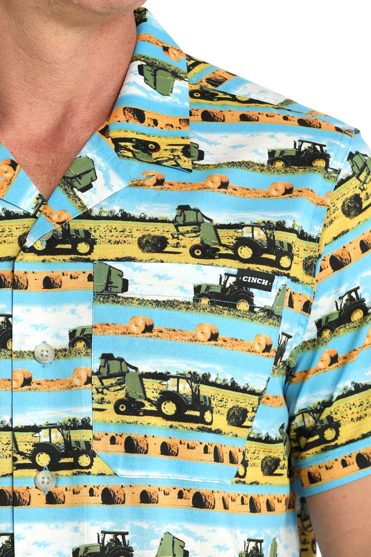 Close-up view of a blue short-sleeve button-down shirt with a multicolor scenic truck and farm print displayed on a white background.