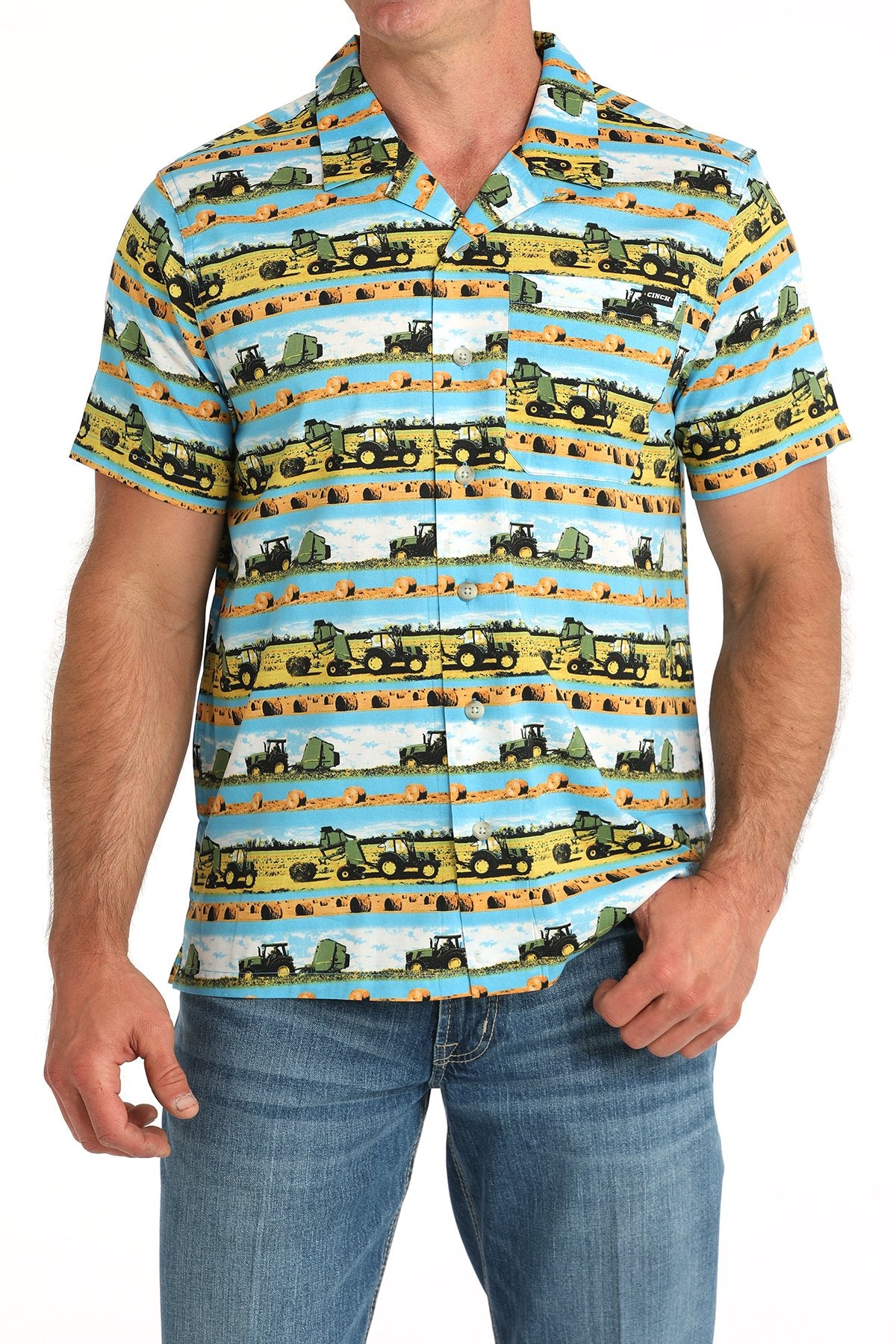 Front view of a man wearing a blue short-sleeve button-down shirt with a multicolor scenic truck and farm print, paired with blue jeans displayed on a white background.