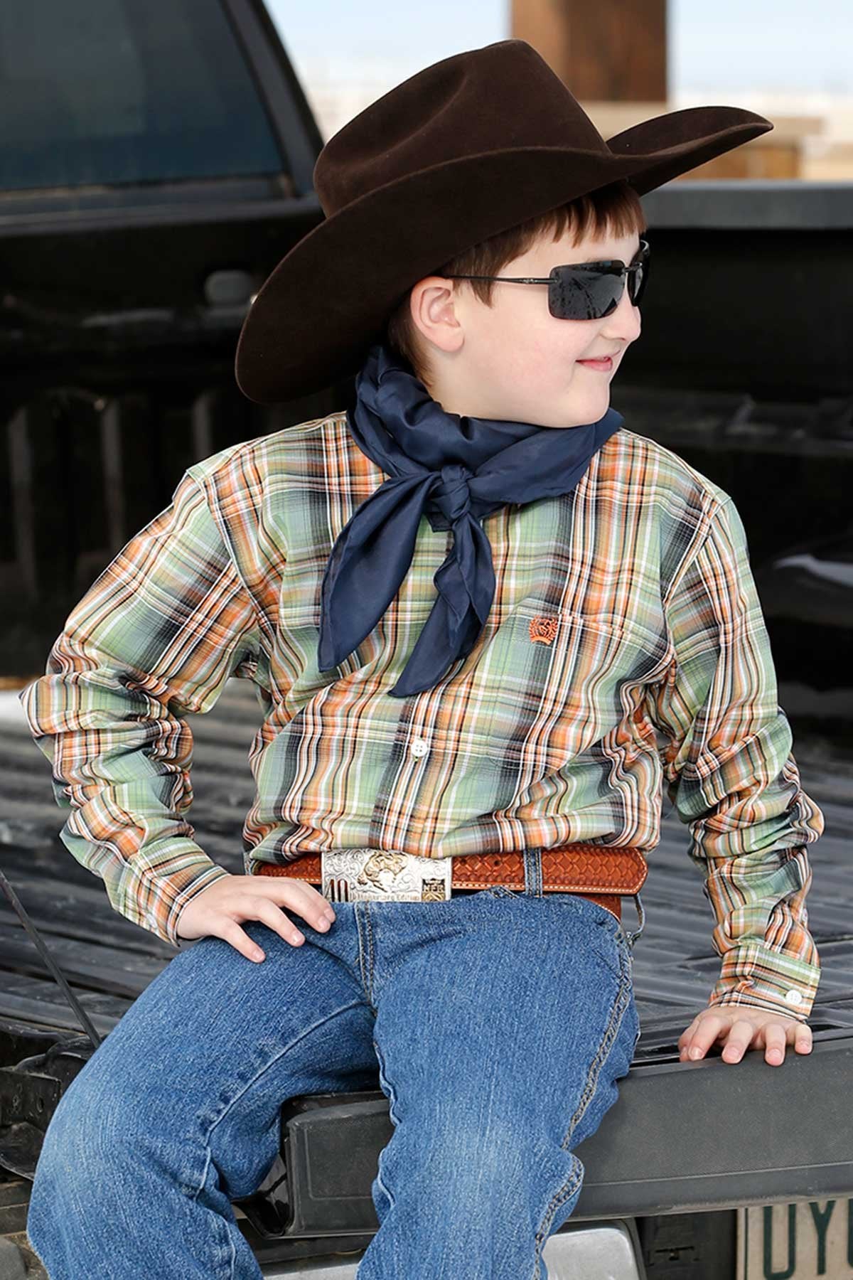 Boy sitting on a truck bed wearing a long sleeve button-down and jeans with a cowboy hat