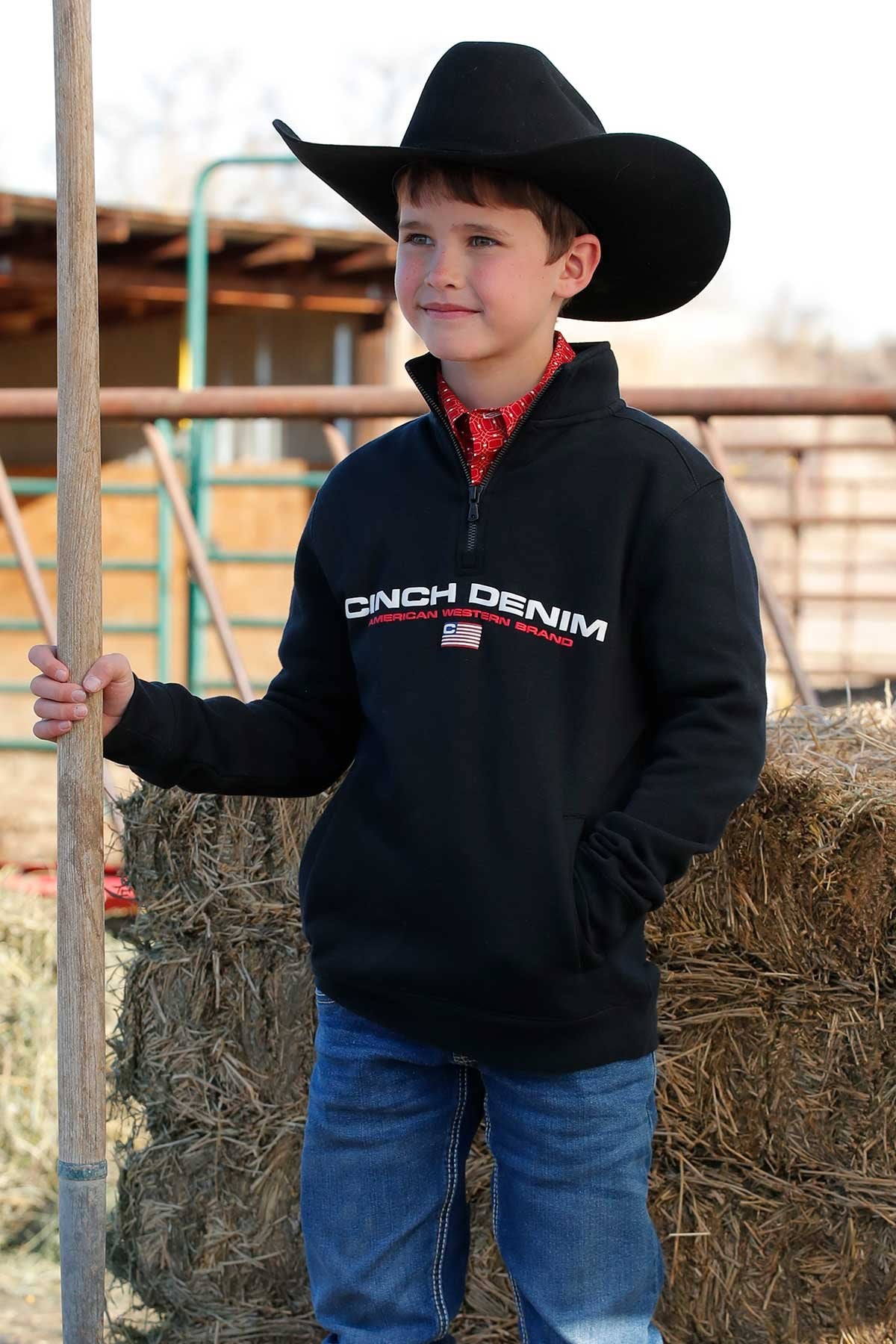 Young boy wearing a black cowboy hat, black quarter-zip pullover with 'Cinch Denim' logo, red patterned shirt underneath, and blue jeans while leaning against hay bales outdoors.
