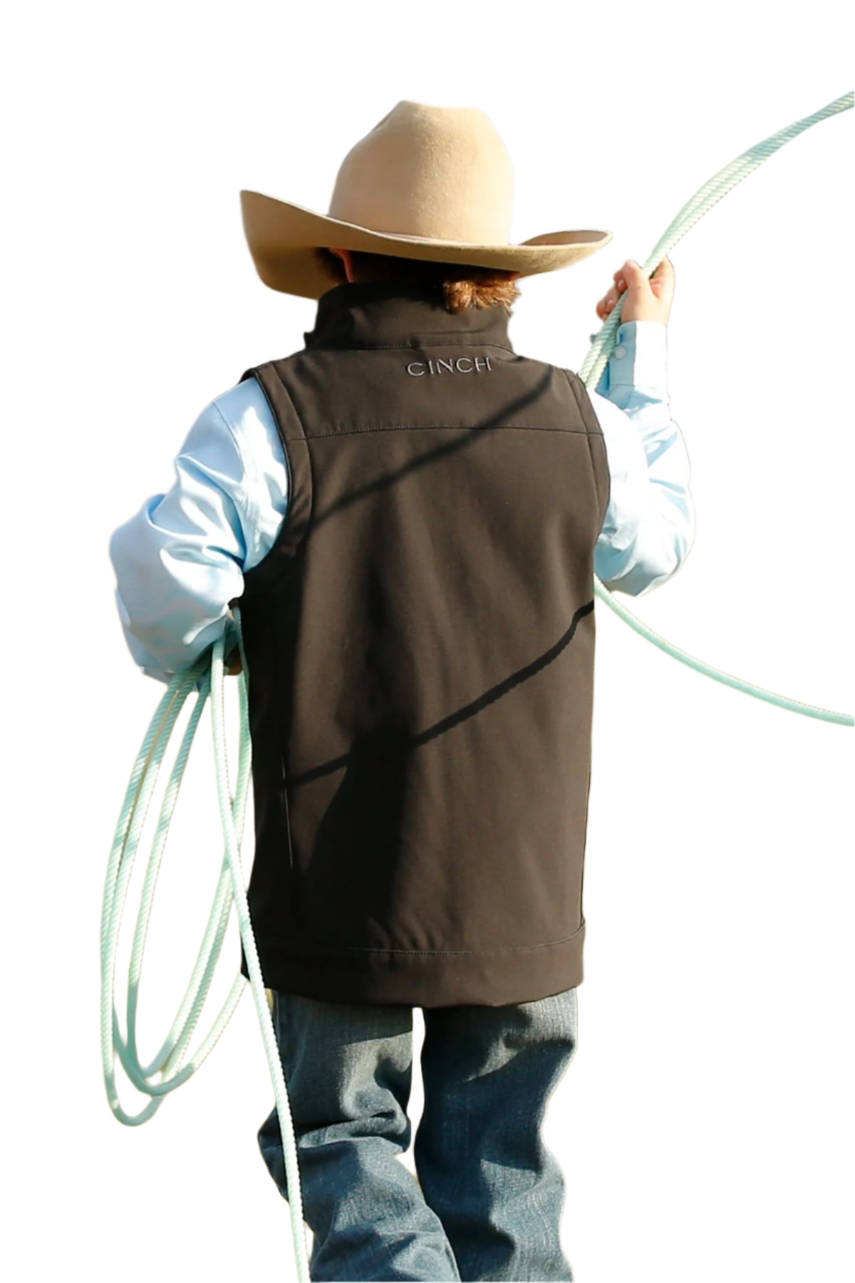 Back view of a boy wearing a black vest over a blue long sleeve shirt and jeans with a cowboy hat holding a rope