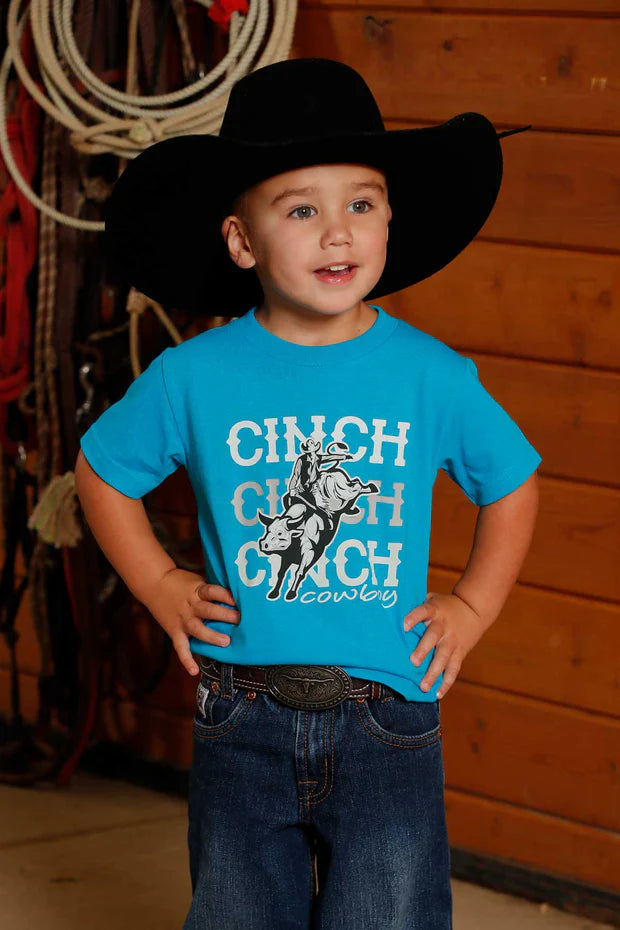 Young boy wearing a teal short sleeve Cinch t-shirt with cowboy bronco rider graphic, jeans, and a cowboy hat, standing indoors against a wooden wall