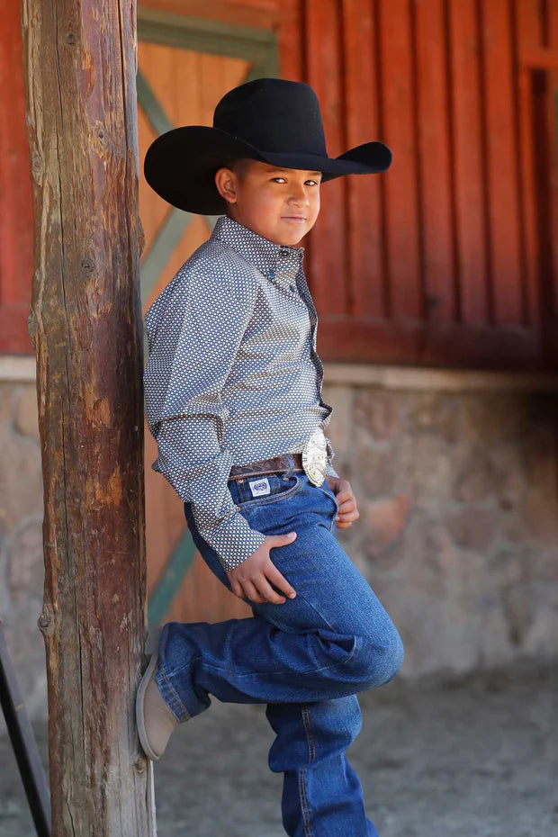 Young boy wearing a teal geometric print long sleeve button-down shirt with jeans, cowboy hat, and belt buckle, leaning against a wooden post