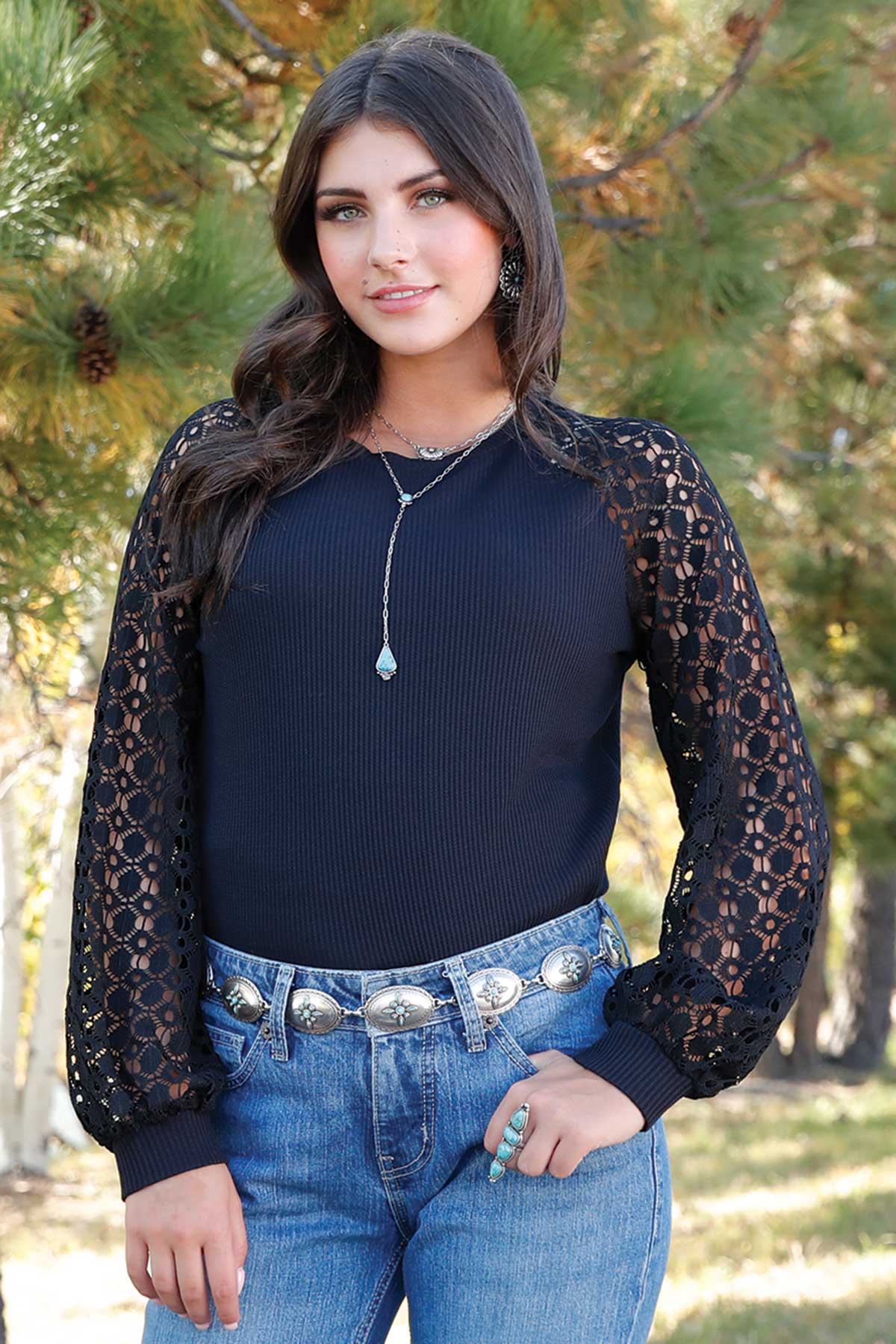 Woman standing outside in front of pine trees, wearing a black long sleeve shirt with contrasting see-through sleeves, paired with jeans and a belt 