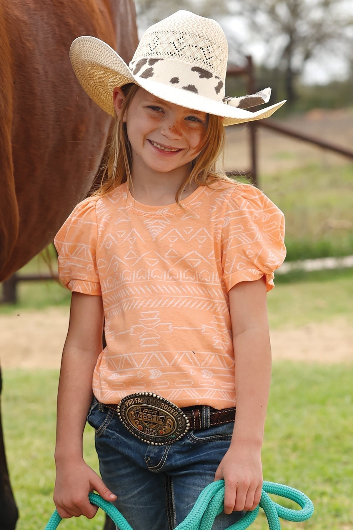 Young girl in a cowboy hat with a Coral Cinch tshirt, standing next to a horse, holding a green rope.
